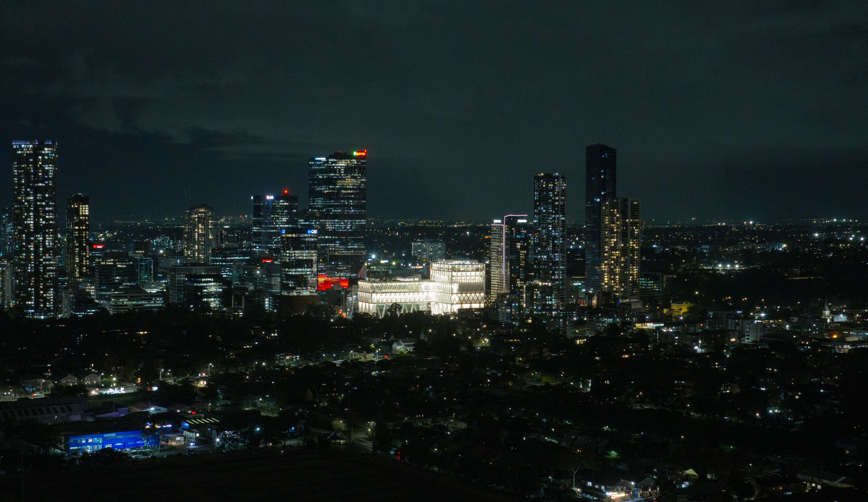 Aerial drone image of a suburban city at night time, focusing on a bright large building (Powerhouse Parramatta) with white beams as its exterior exoskeleton.