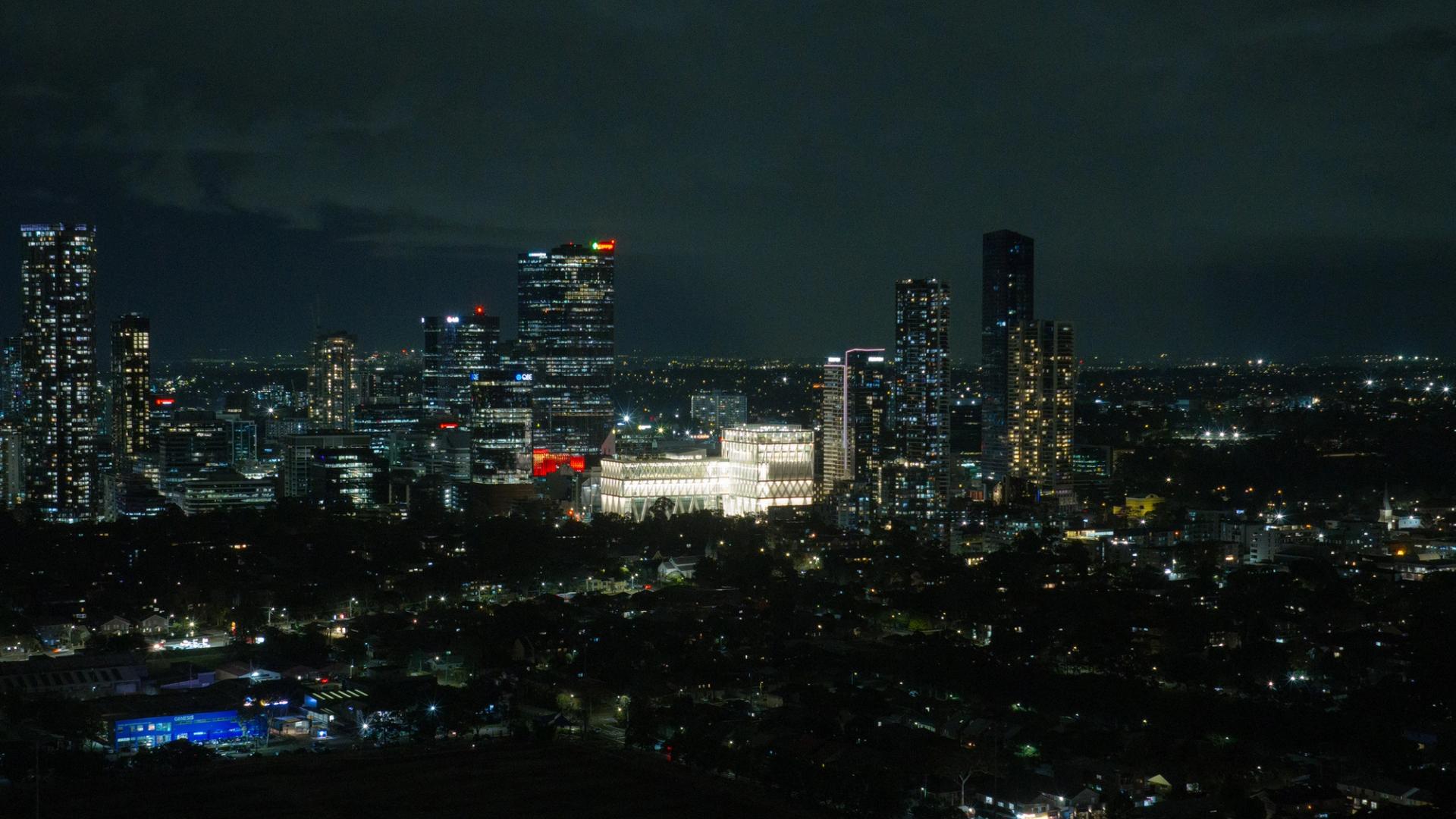 Aerial drone image of a suburban city at night time, focusing on a bright large building (Powerhouse Parramatta) with white beams as its exterior exoskeleton.