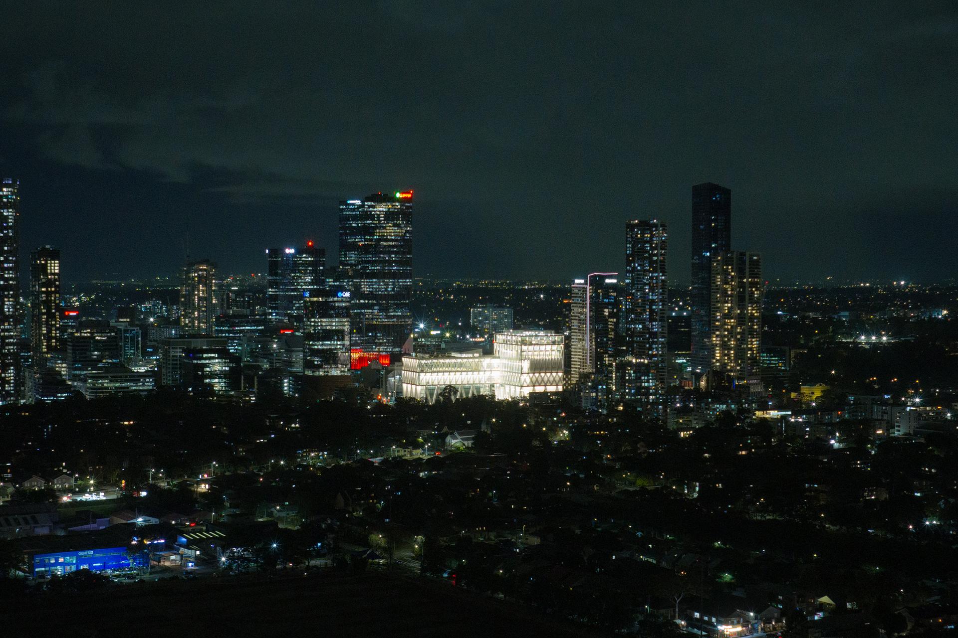 Aerial drone image of a suburban city at night time, focusing on a bright large building (Powerhouse Parramatta) with white beams as its exterior exoskeleton.