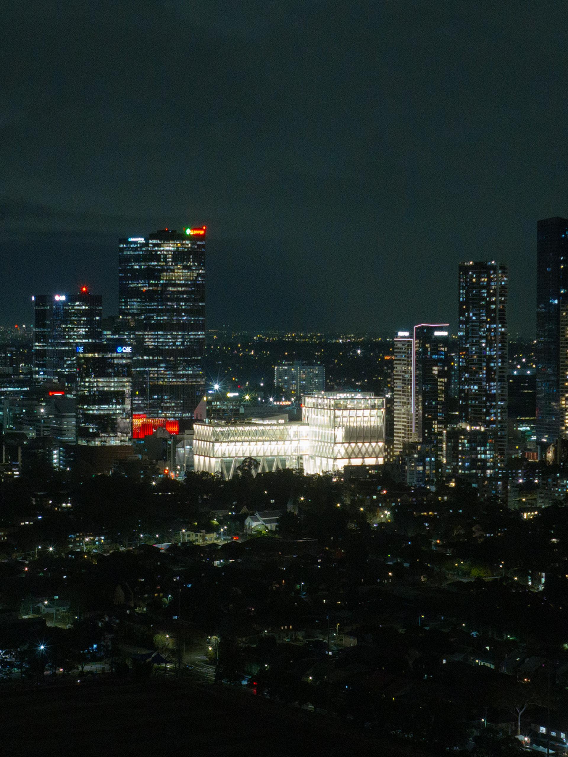 Aerial drone image of a suburban city at night time, focusing on a bright large building (Powerhouse Parramatta) with white beams as its exterior exoskeleton.