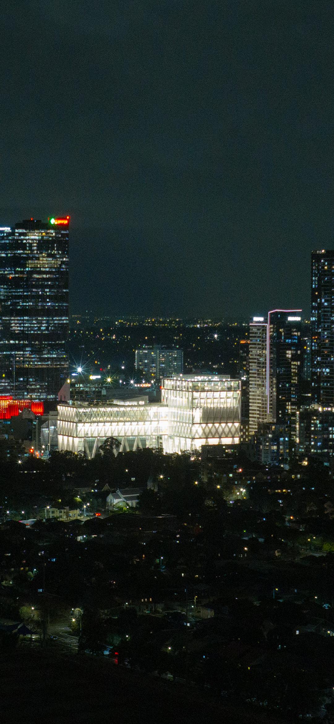Aerial drone image of a suburban city at night time, focusing on a bright large building (Powerhouse Parramatta) with white beams as its exterior exoskeleton.