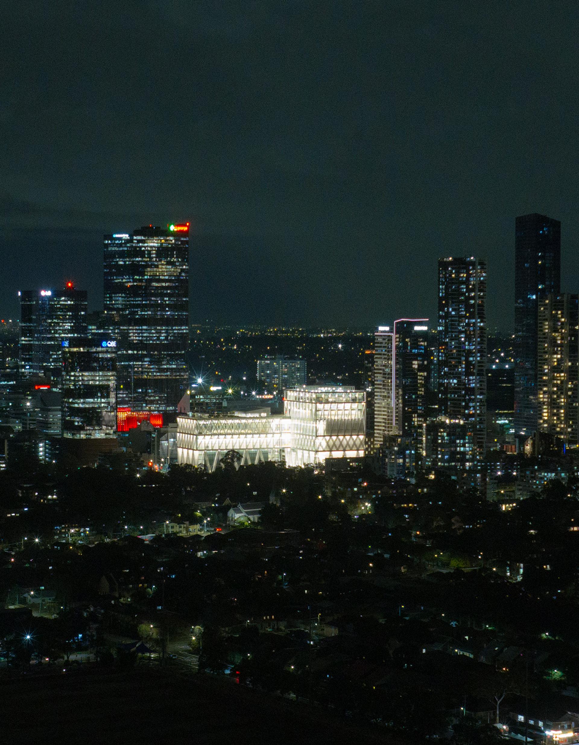 Aerial drone image of a suburban city at night time, focusing on a bright large building (Powerhouse Parramatta) with white beams as its exterior exoskeleton.
