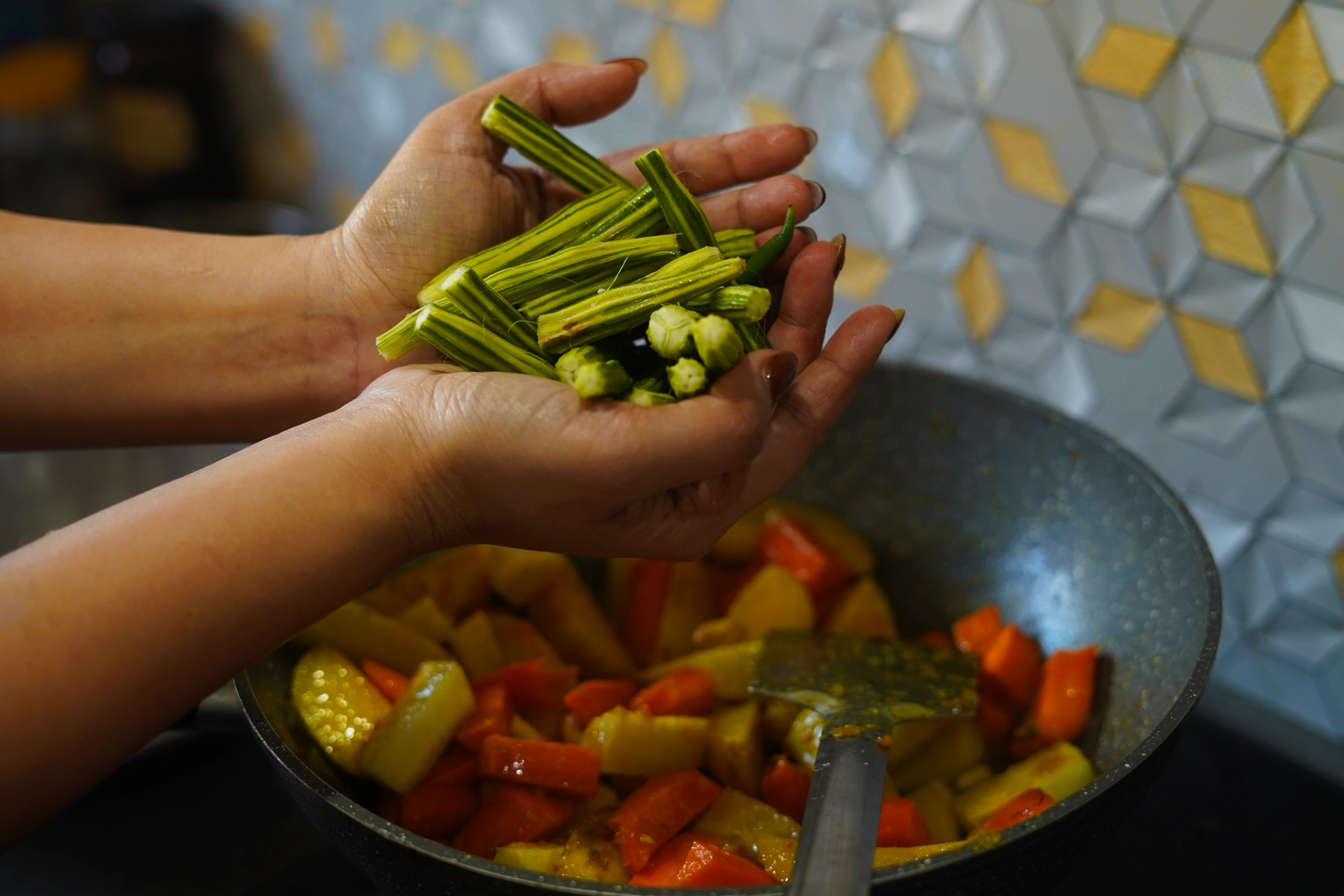 In the foreground, two brown hands hold green moringa pods. In the background, sticks of potato and carrot are cooking in a large pan.