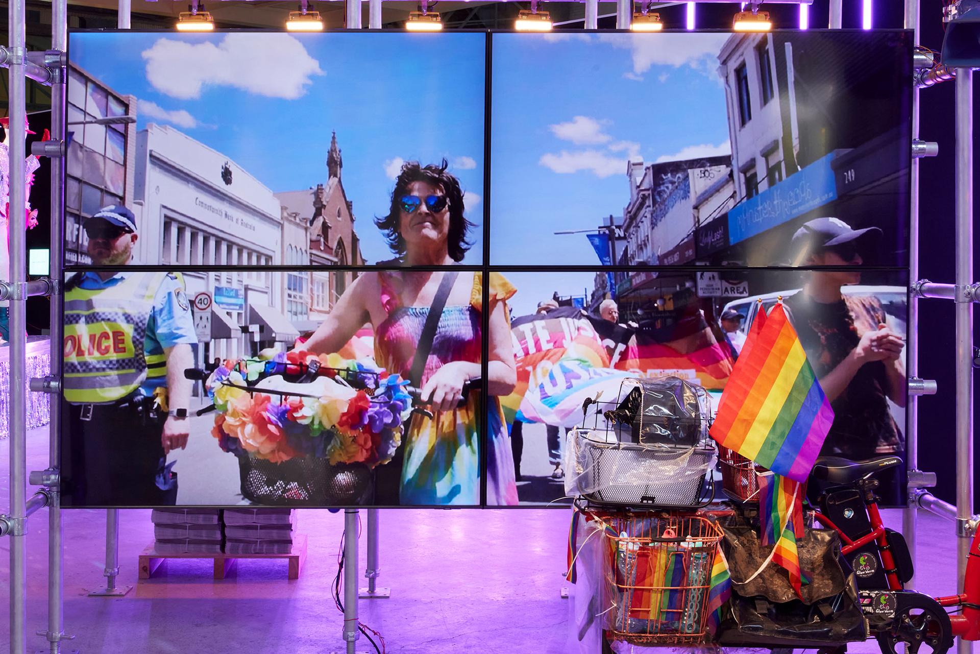 A screen in an exhibition space. On the screen is Norrie, a non-binary person wearing a shirred-top rainbow coloured dress, aviator sunglasses with blue lenses and a cross body bag draped over their left shoulder. They have mid length brown hair. Norrie is pushing a bicycle that has a rainbow-coloured lei wrapped around the front basket. To Norrie’s right is a police officer. Behind Norrie protestors are marching with a large, coloured banner.