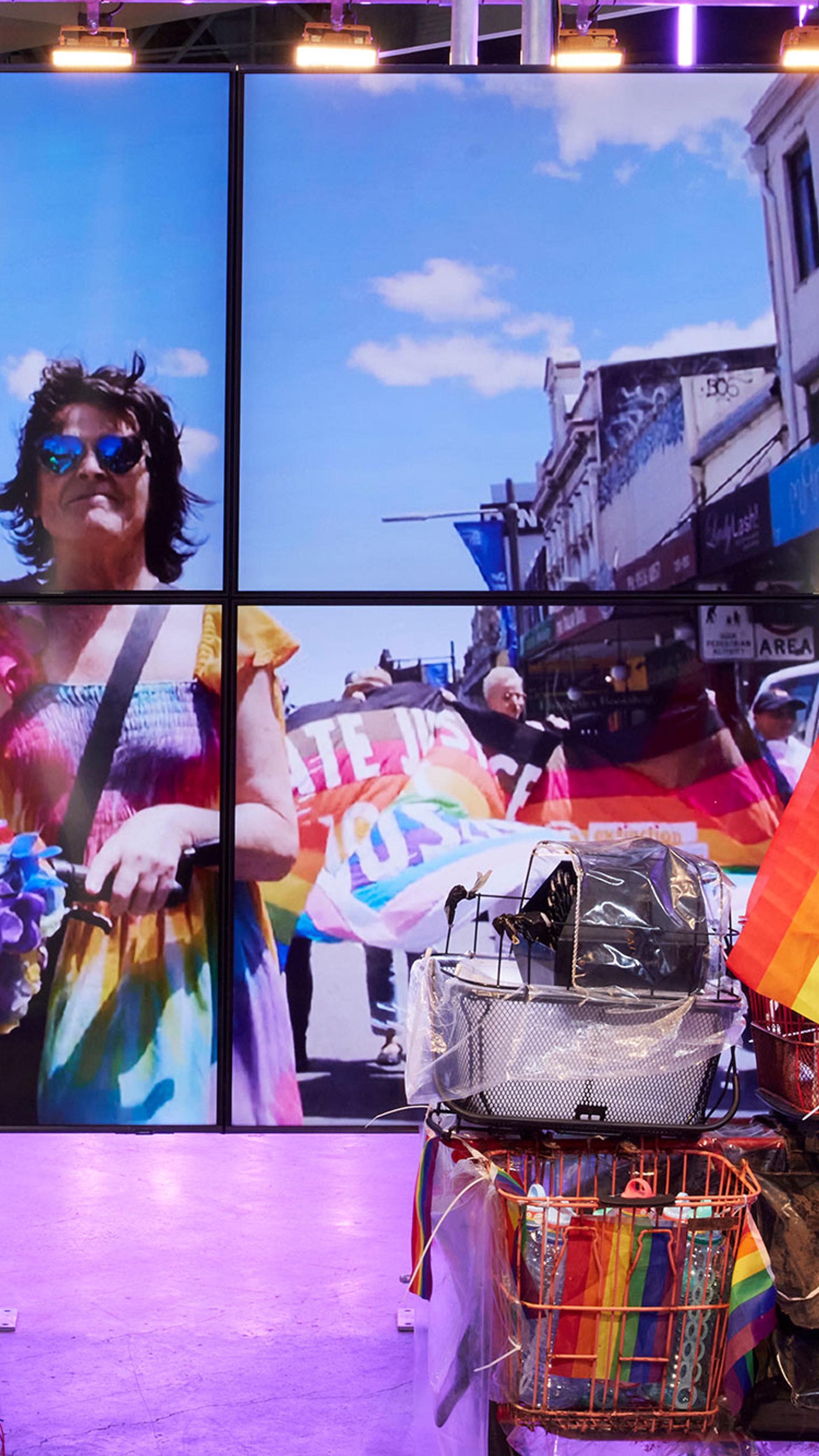 A screen in an exhibition space. On the screen is Norrie, a non-binary person wearing a shirred-top rainbow coloured dress, aviator sunglasses with blue lenses and a cross body bag draped over their left shoulder. They have mid length brown hair. Norrie is pushing a bicycle that has a rainbow-coloured lei wrapped around the front basket. To Norrie’s right is a police officer. Behind Norrie protestors are marching with a large, coloured banner.