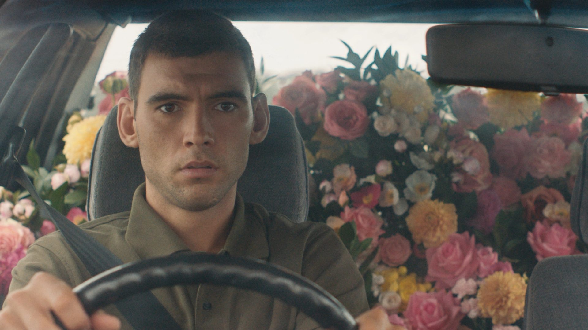 A still of a film, featuring an actor in the front seat of a car with the backseats filled with pink, yellow and white flowers.