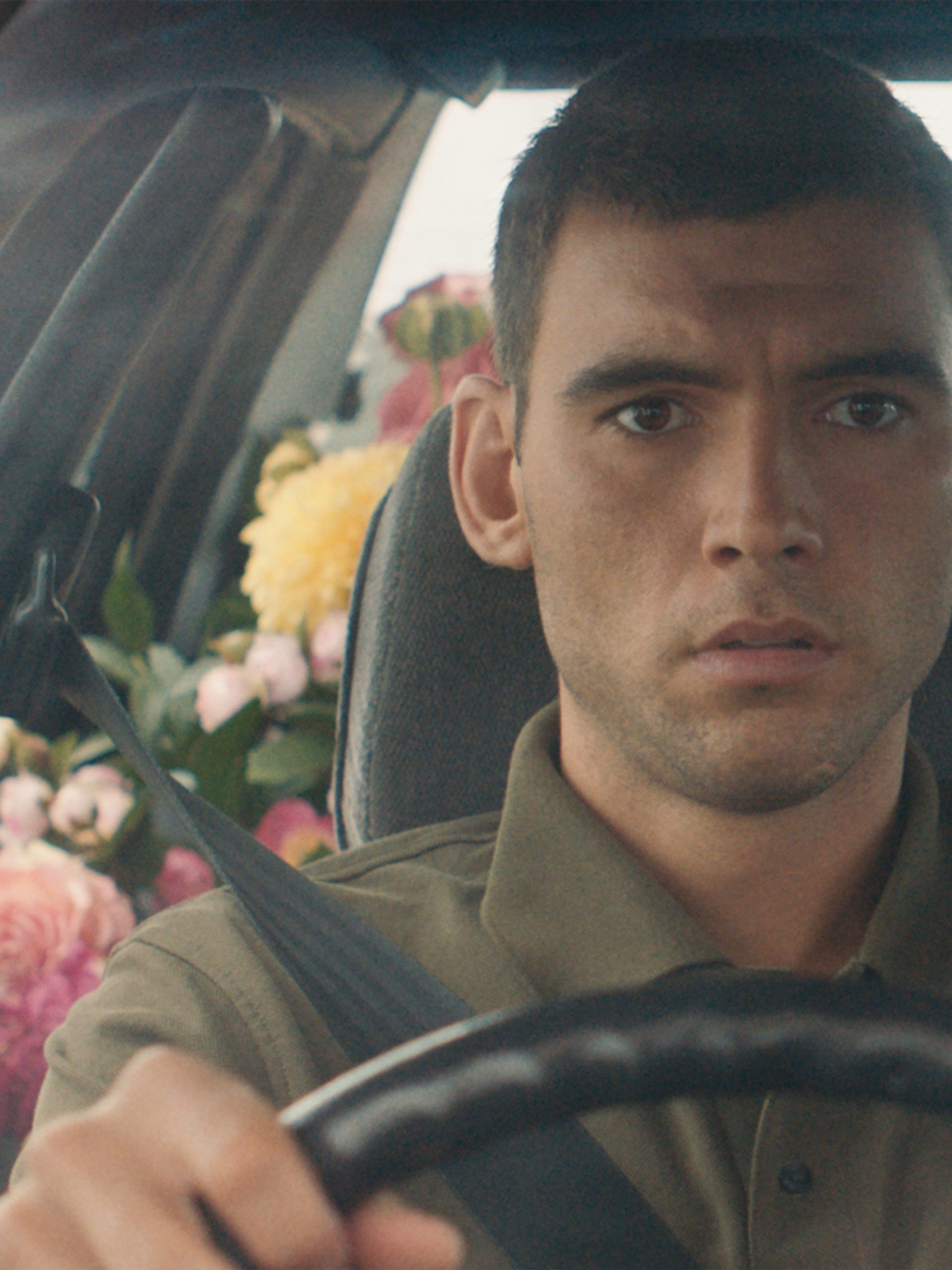 A still of a film, featuring an actor in the front seat of a car with the backseats filled with pink, yellow and white flowers.