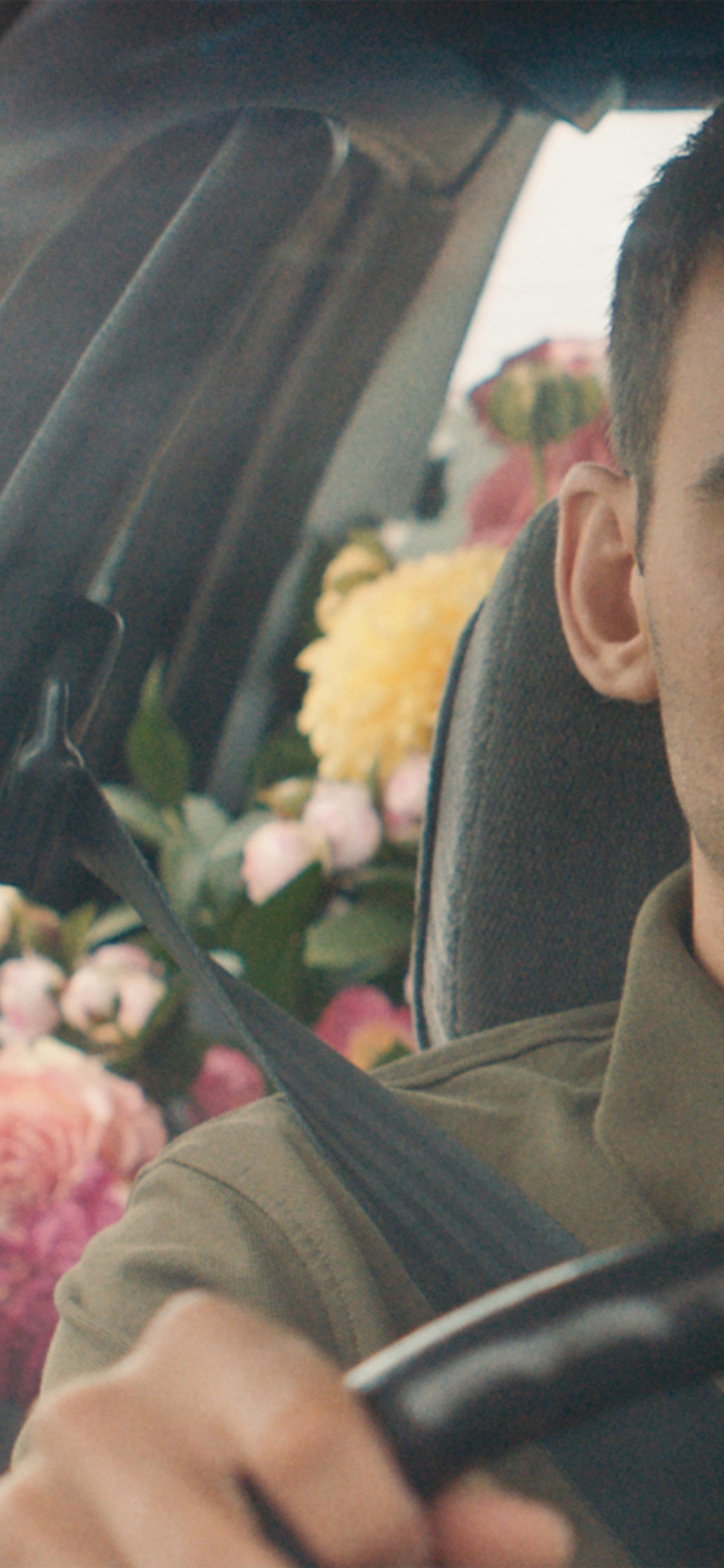 A still of a film, featuring an actor in the front seat of a car with the backseats filled with pink, yellow and white flowers.