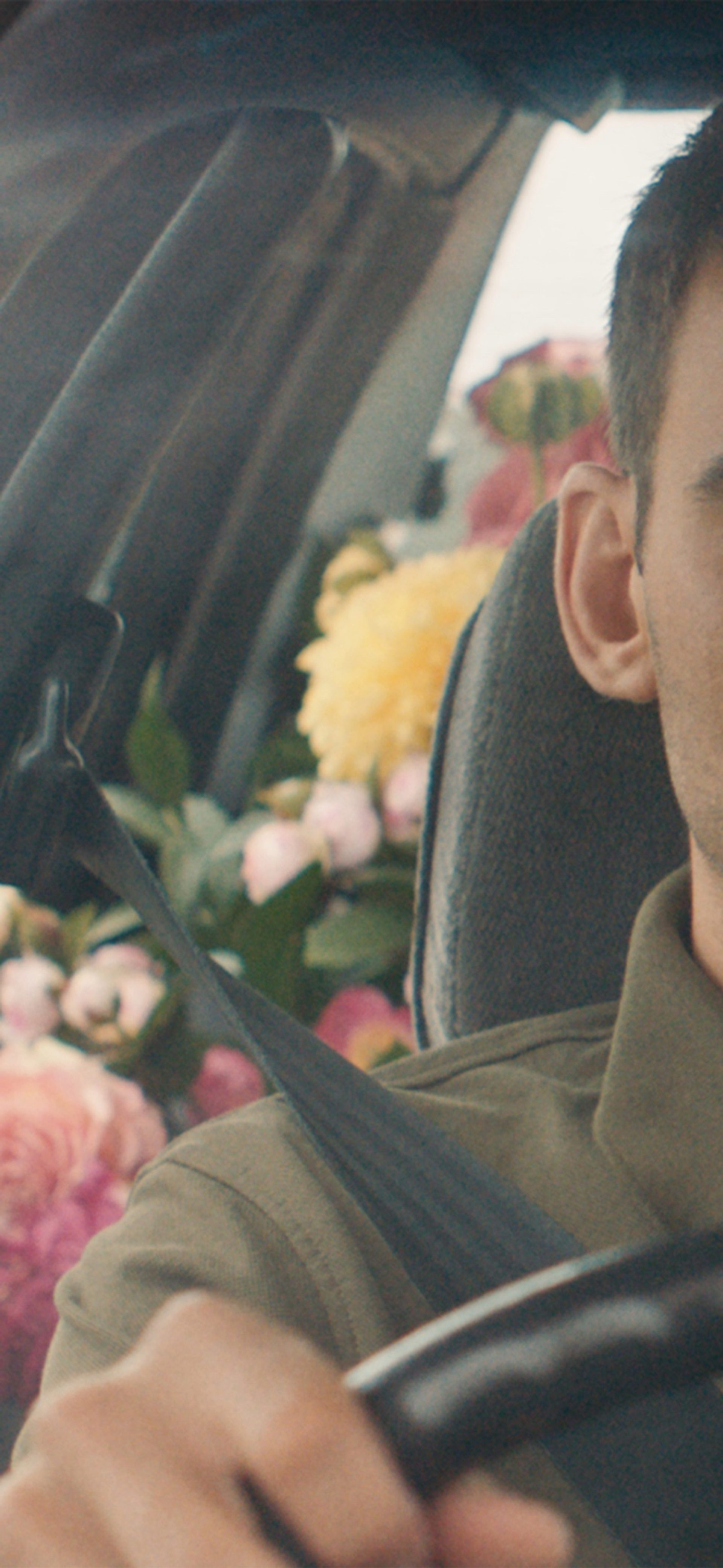 A still of a film, featuring an actor in the front seat of a car with the backseats filled with pink, yellow and white flowers.