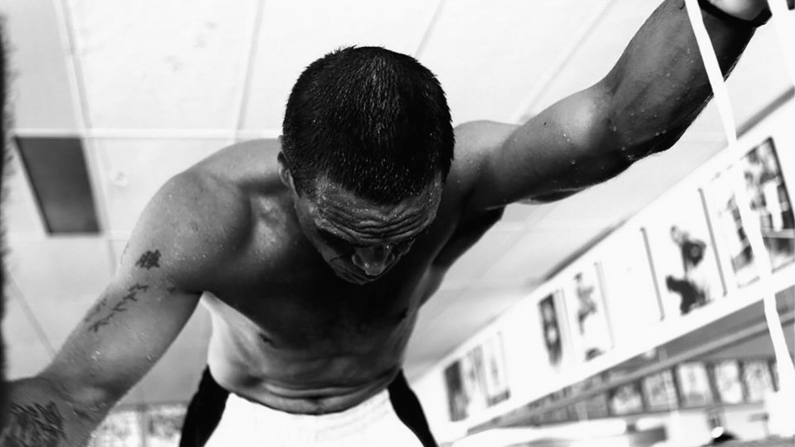 A black and white photograph of a man stretched across the camera, shirtless. His right arm is tattooed and visible.