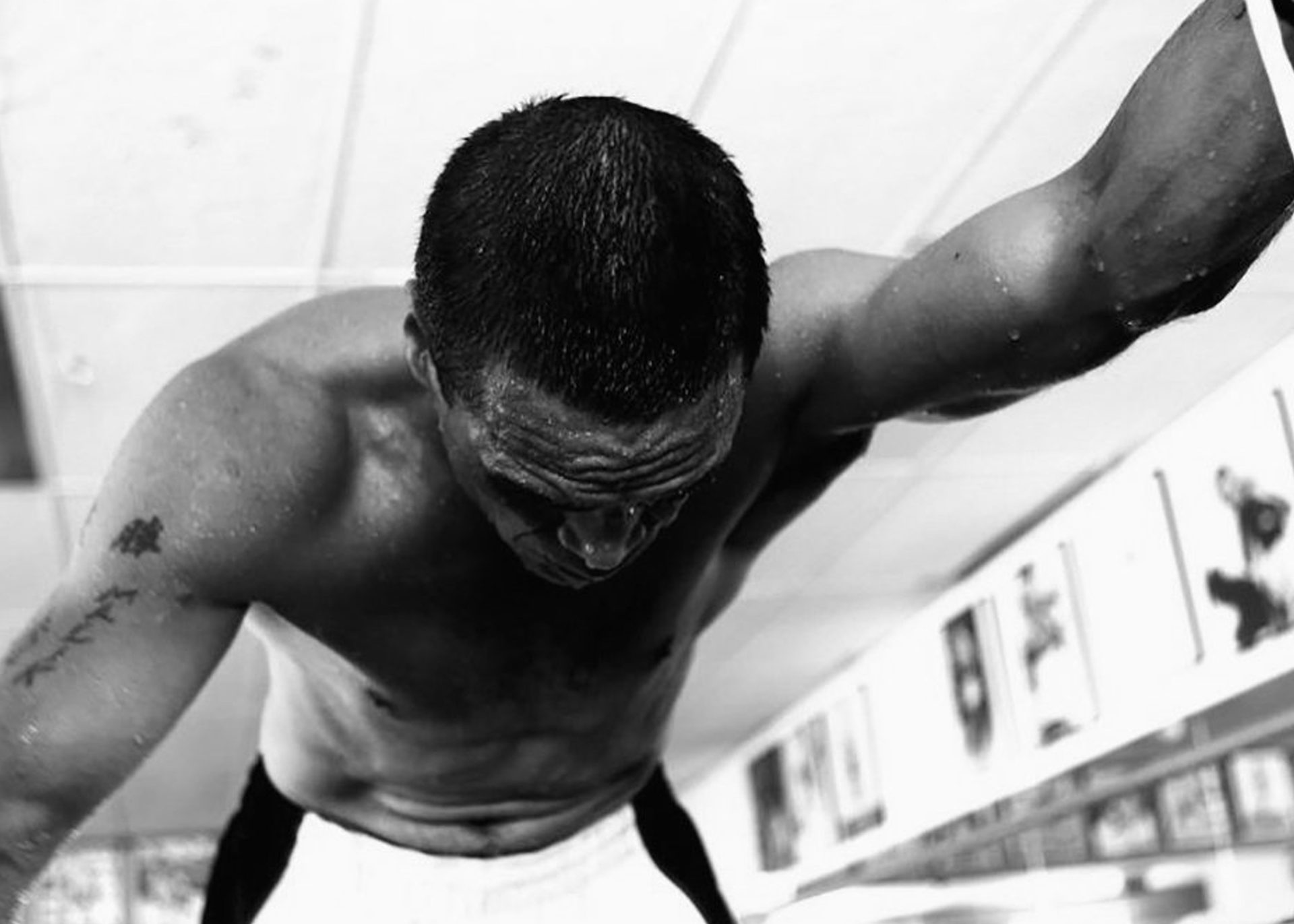 A black and white photograph of a man stretched across the camera, shirtless. His right arm is tattooed and visible.