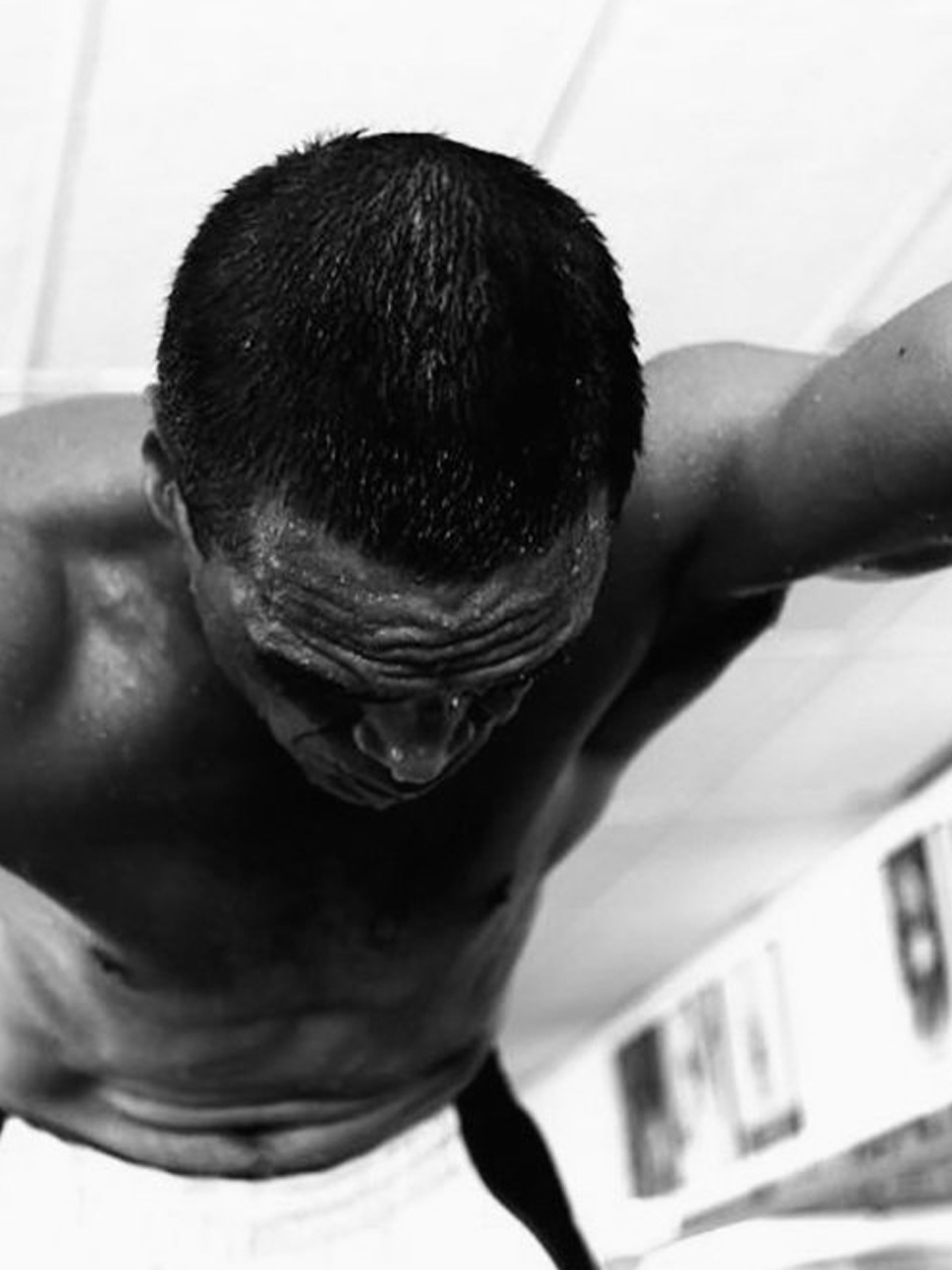 A black and white photograph of a man stretched across the camera, shirtless. His right arm is tattooed and visible.