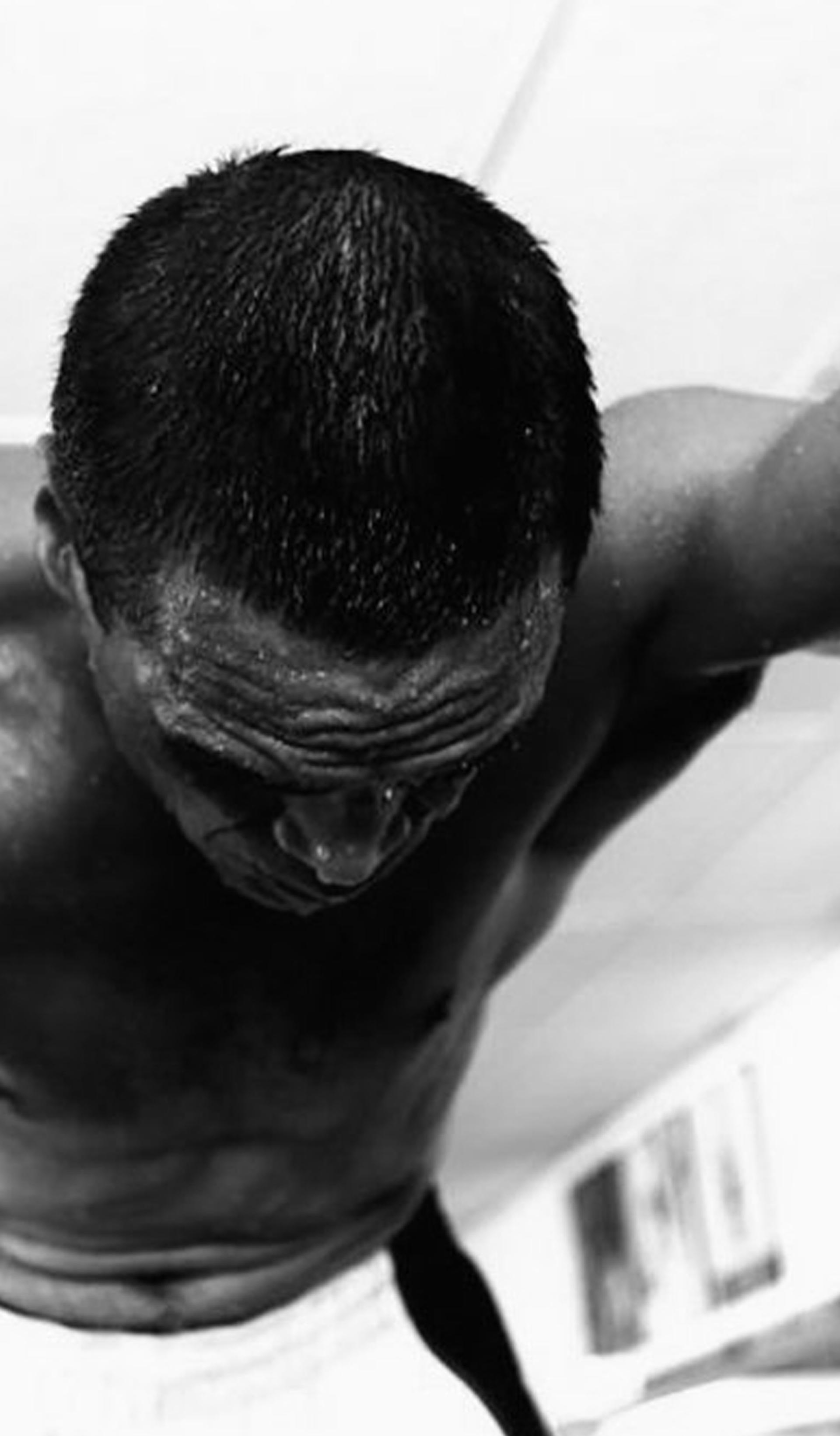 A black and white photograph of a man stretched across the camera, shirtless. His right arm is tattooed and visible.