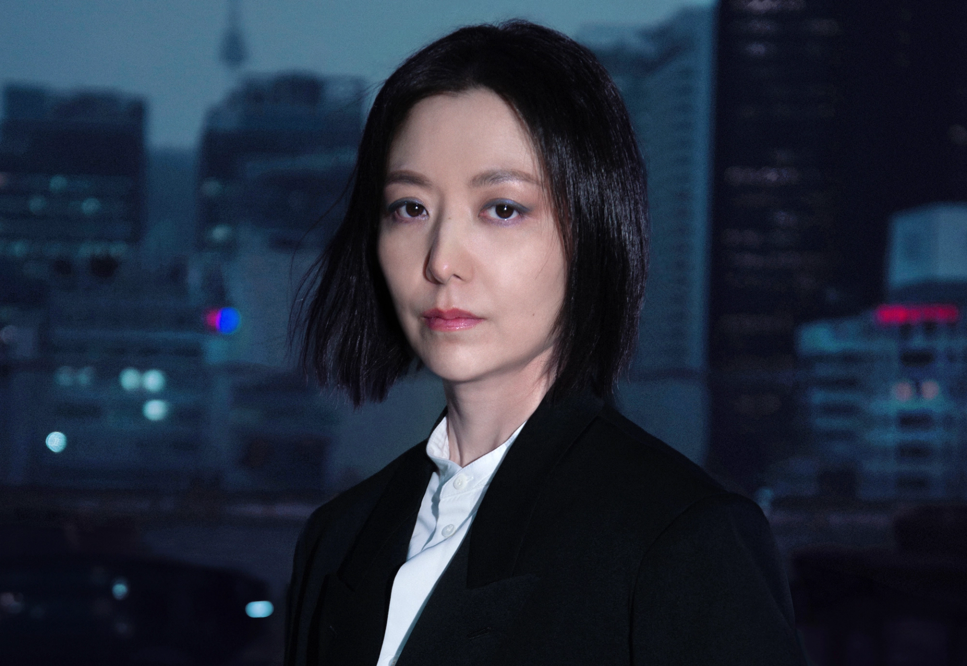 The artist Ayoung Kim standing in front of a city skyline, the light fading. She stares straight at the camera.
