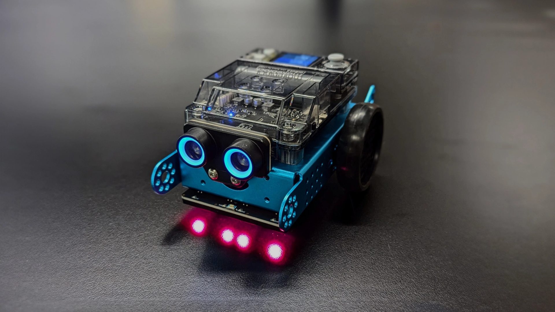 A small car-shaped blue robot with blue sensor eyes and four red headlights sits on a dark grey background.