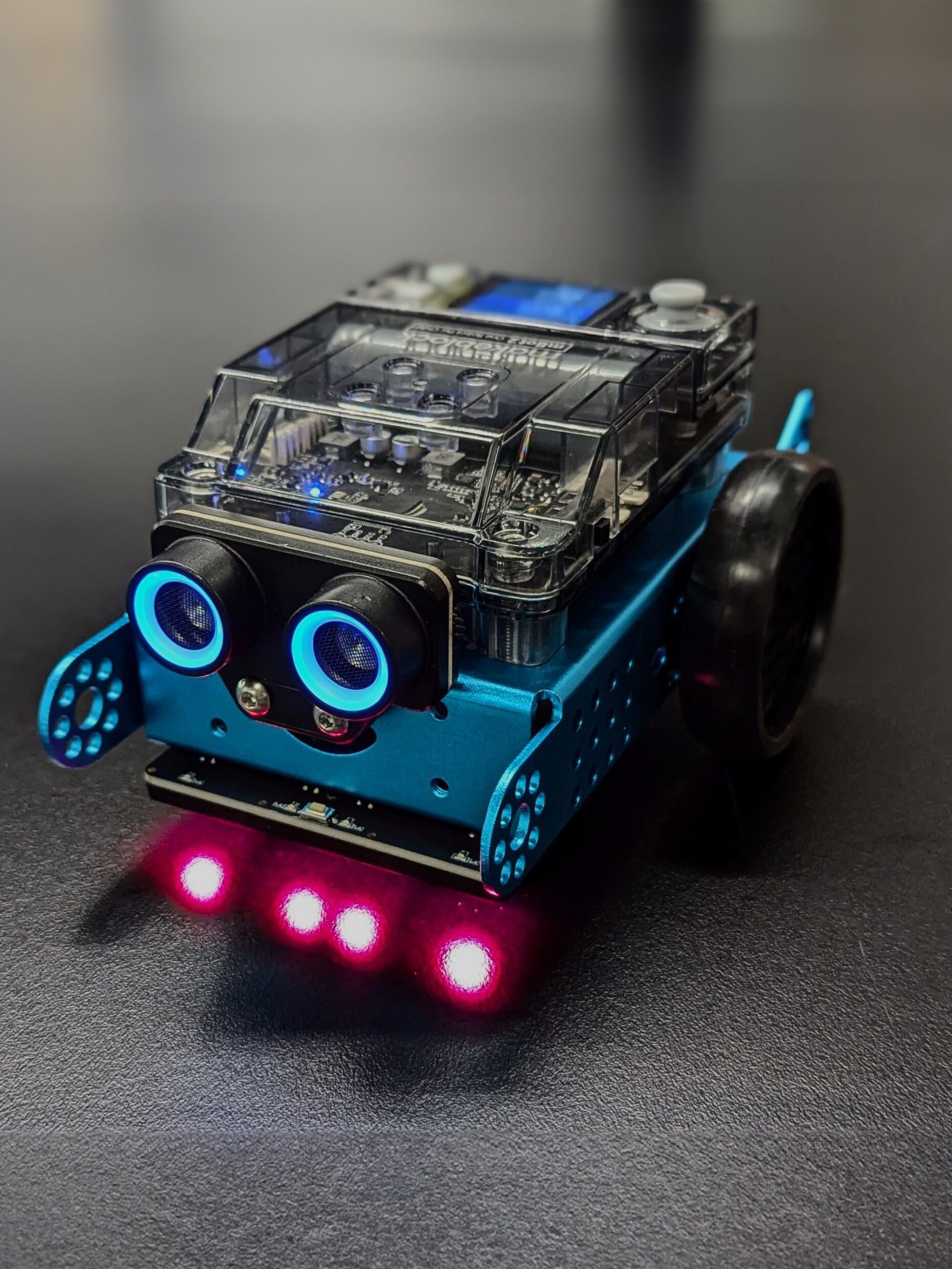 A small car-shaped blue robot with blue sensor eyes and four red headlights sits on a dark grey background.