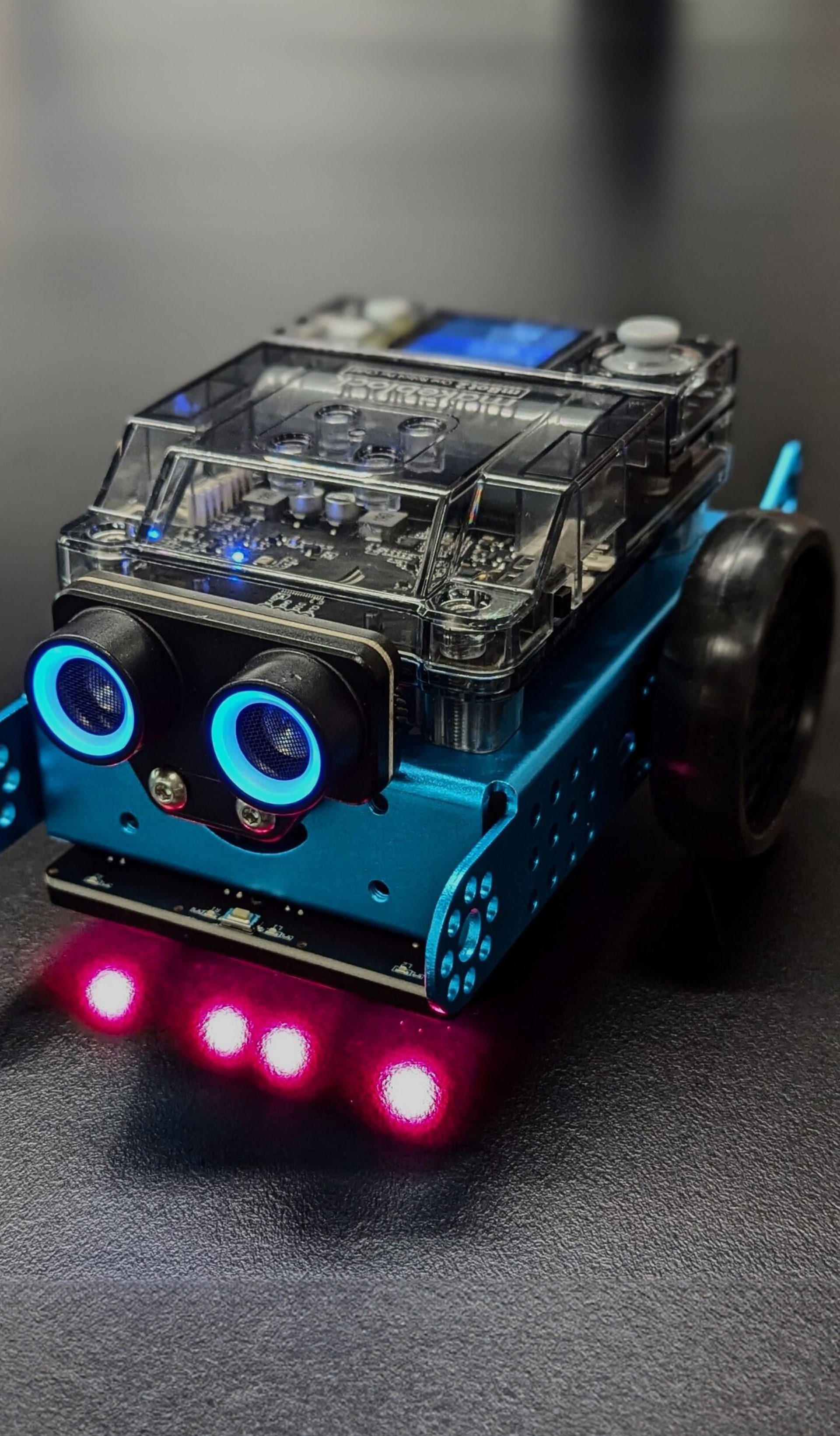 A small car-shaped blue robot with blue sensor eyes and four red headlights sits on a dark grey background.