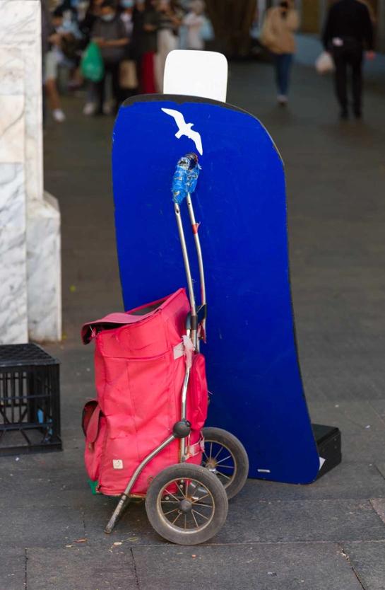 A red shopping trolley with two wheels leaning up against a blue sign with a white bird on it.