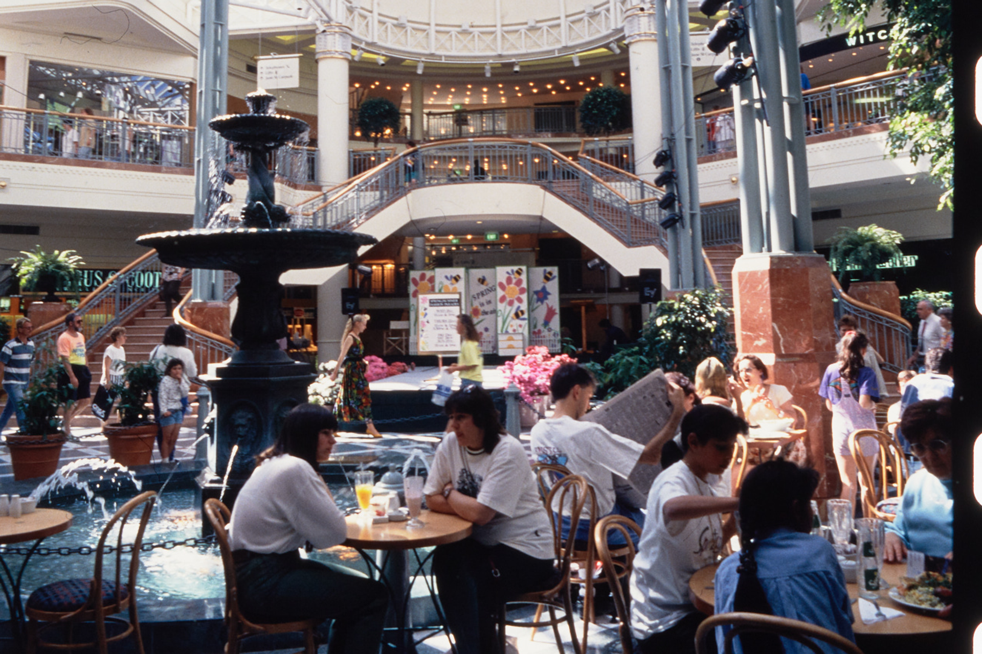 A photograph of the busy Food court at Penrith Plaza, cafe tables filled with guests surround the centre fountain, with a two-way staircase in the background.