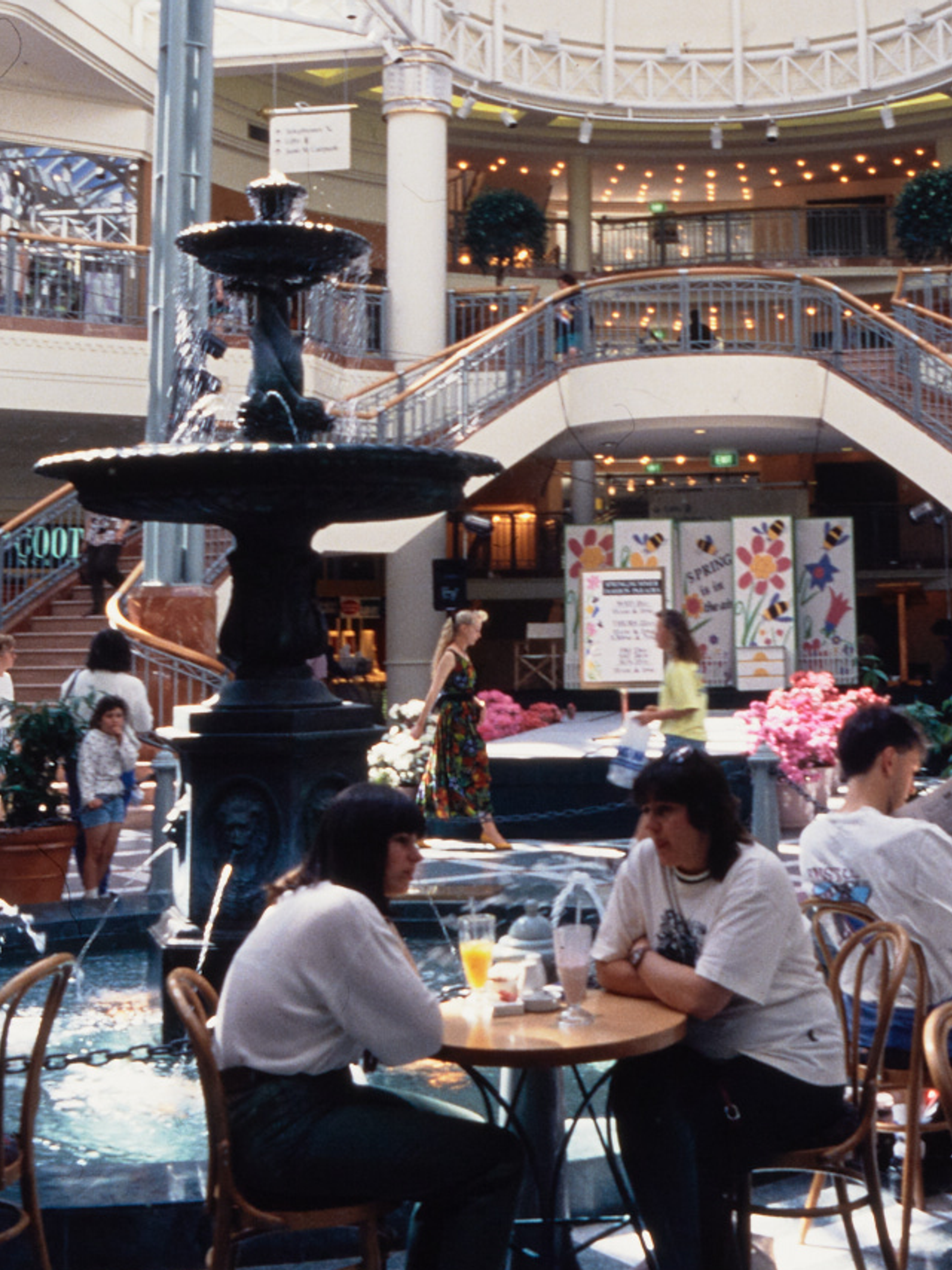 A photograph of the busy Food court at Penrith Plaza, cafe tables filled with guests surround the centre fountain, with a two-way staircase in the background.