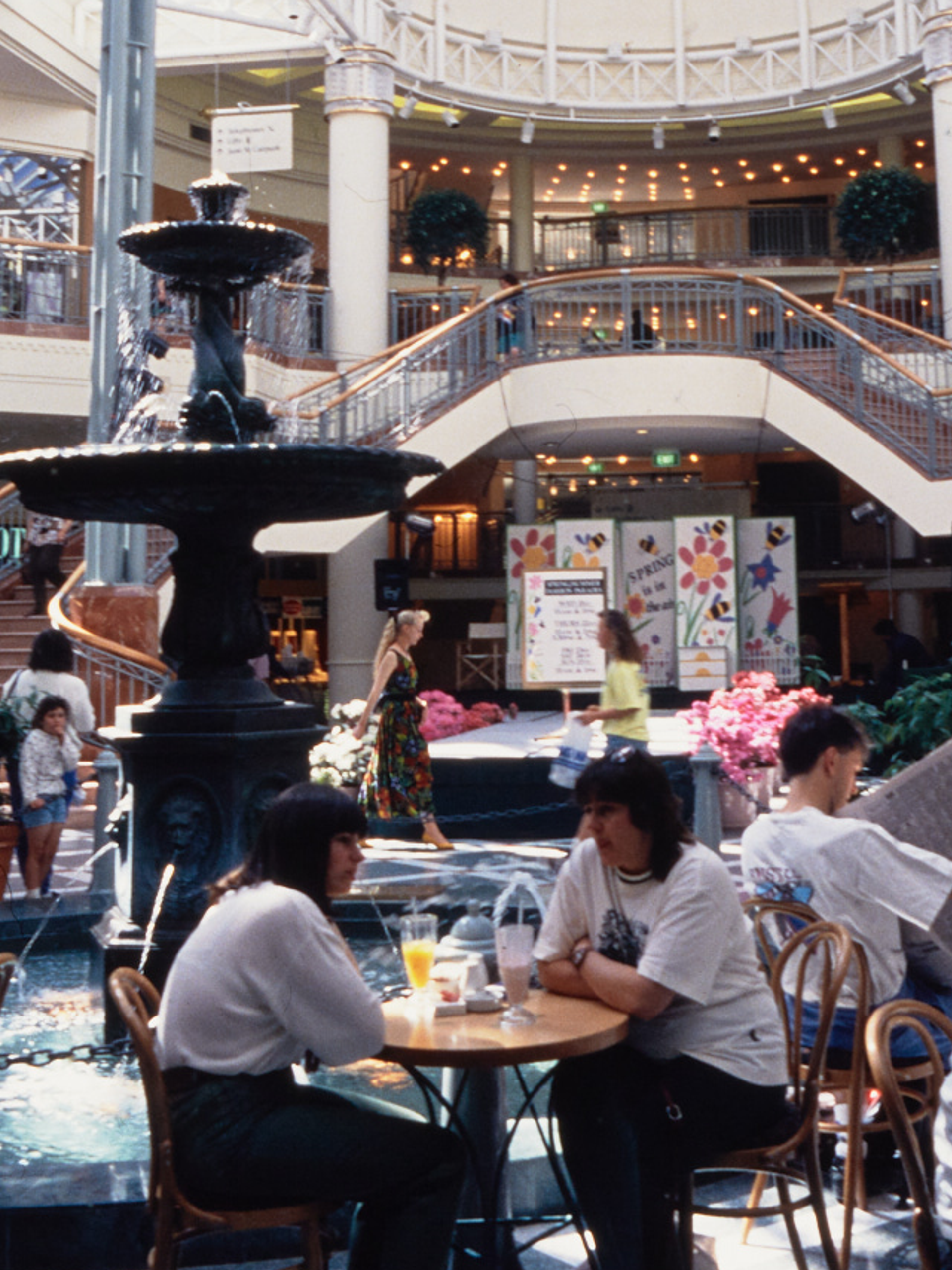 A photograph of the busy Food court at Penrith Plaza, cafe tables filled with guests surround the centre fountain, with a two-way staircase in the background.
