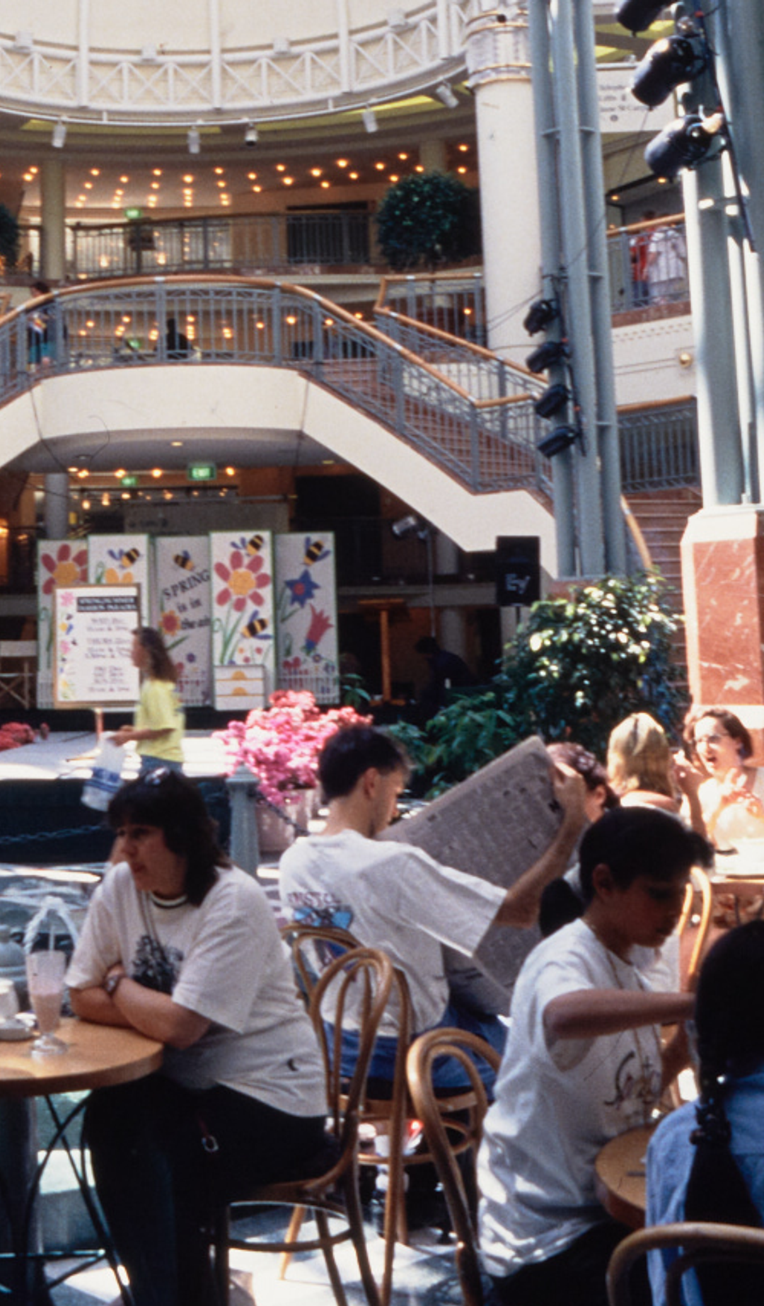A photograph of the busy Food court at Penrith Plaza, cafe tables filled with guests surround the centre fountain, with a two-way staircase in the background.
