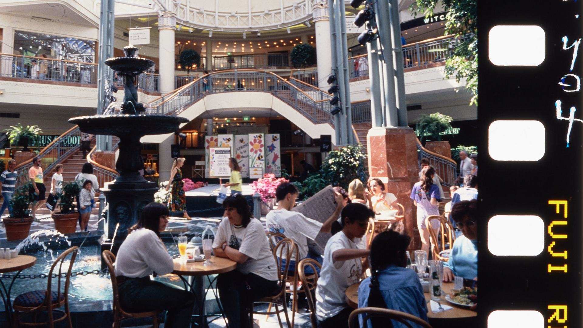 A photograph of the busy Food court at Penrith Plaza, cafe tables filled with guests surround the centre fountain, with a two-way staircase in the background.