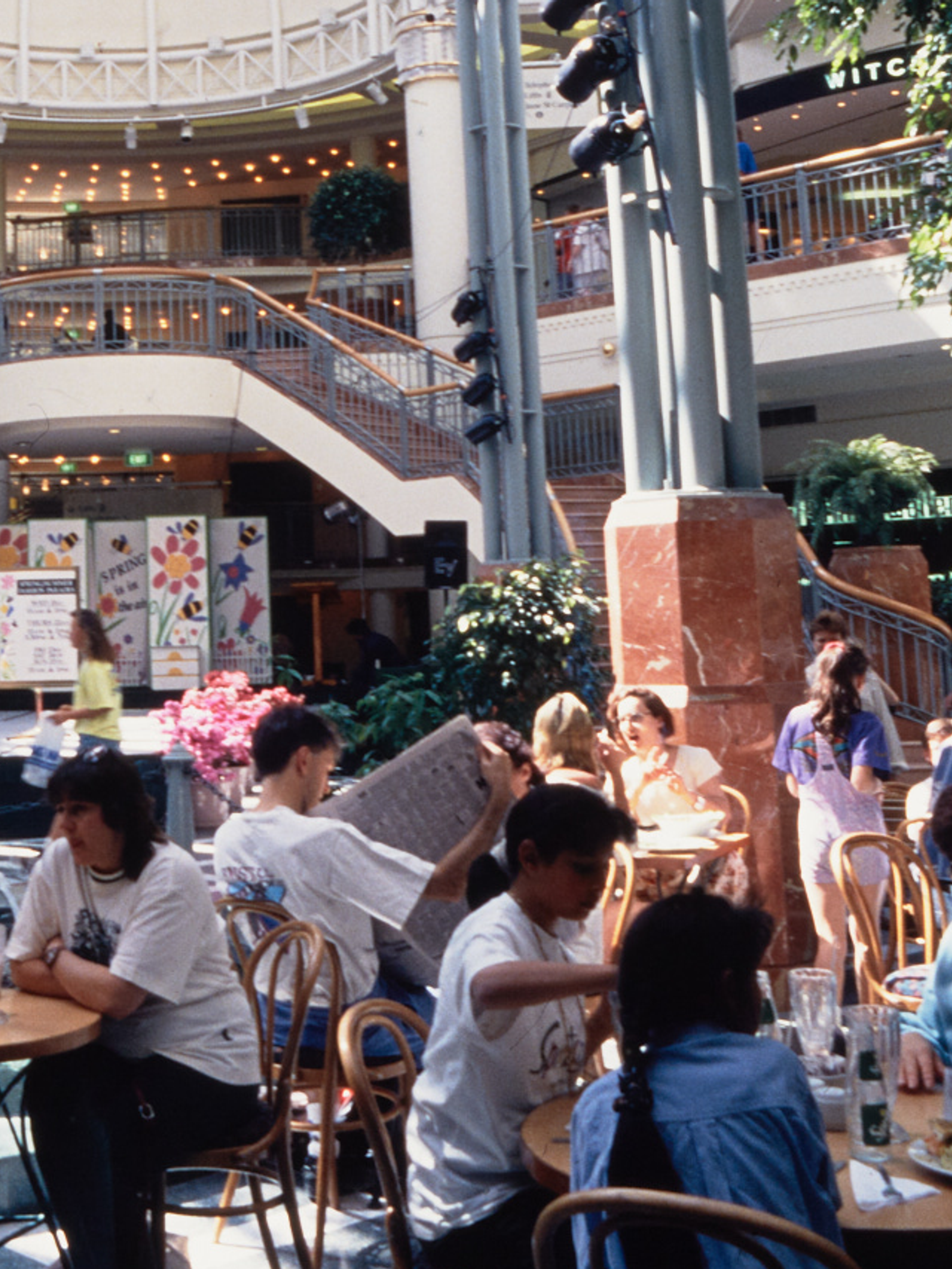 A photograph of the busy Food court at Penrith Plaza, cafe tables filled with guests surround the centre fountain, with a two-way staircase in the background.
