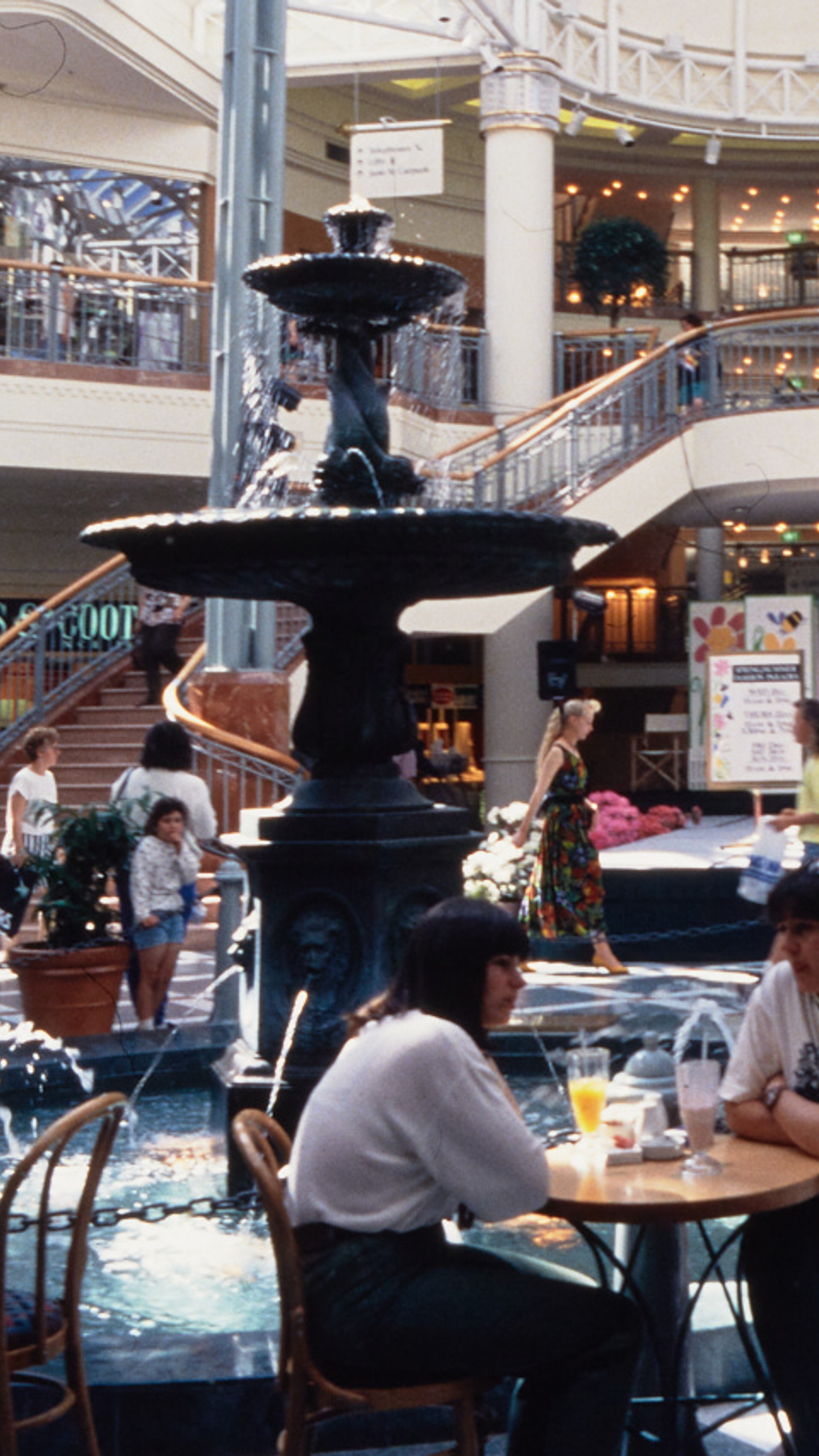 A photograph of the busy Food court at Penrith Plaza, cafe tables filled with guests surround the centre fountain, with a two-way staircase in the background.