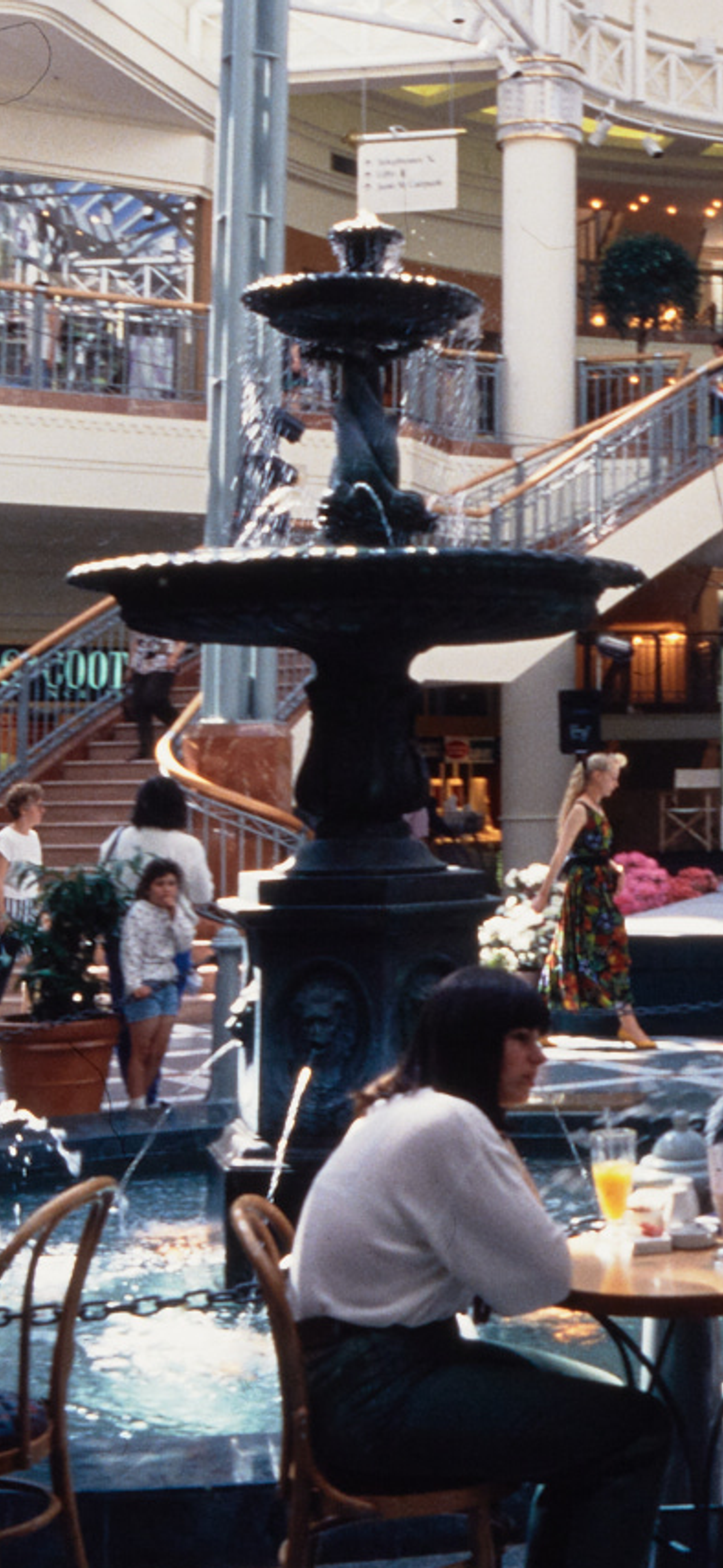 A photograph of the busy Food court at Penrith Plaza, cafe tables filled with guests surround the centre fountain, with a two-way staircase in the background.