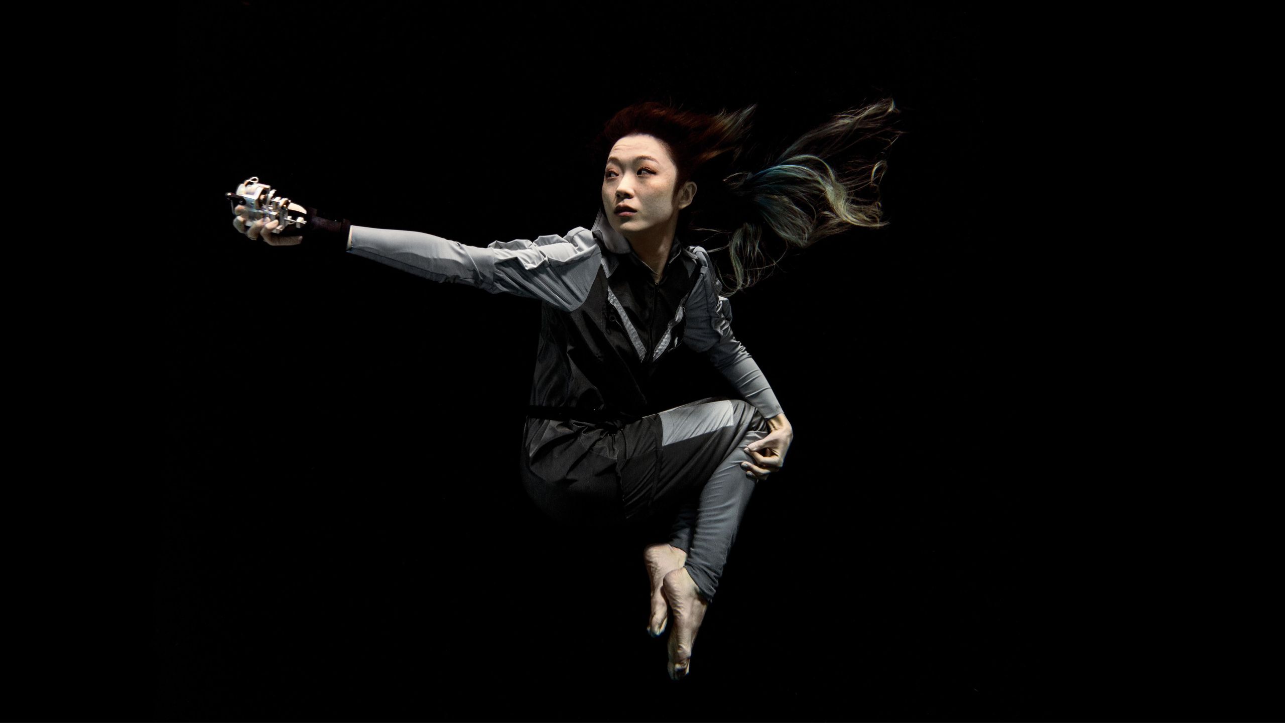 Artist Xin Liu floating with arm outstretched against a black background. She wears a full-length grey body suit with long sleeves with bare feet and hands..