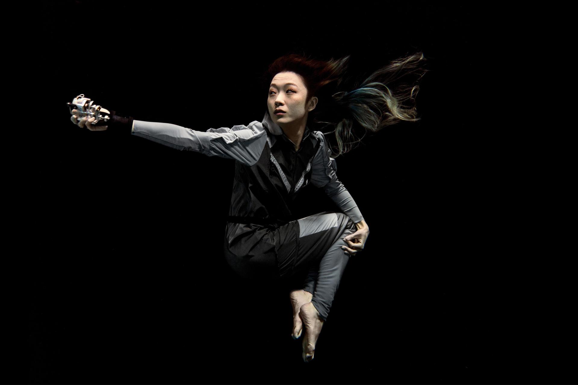 Artist Xin Liu floating with arm outstretched against a black background. She wears a full-length grey body suit with long sleeves with bare feet and hands..