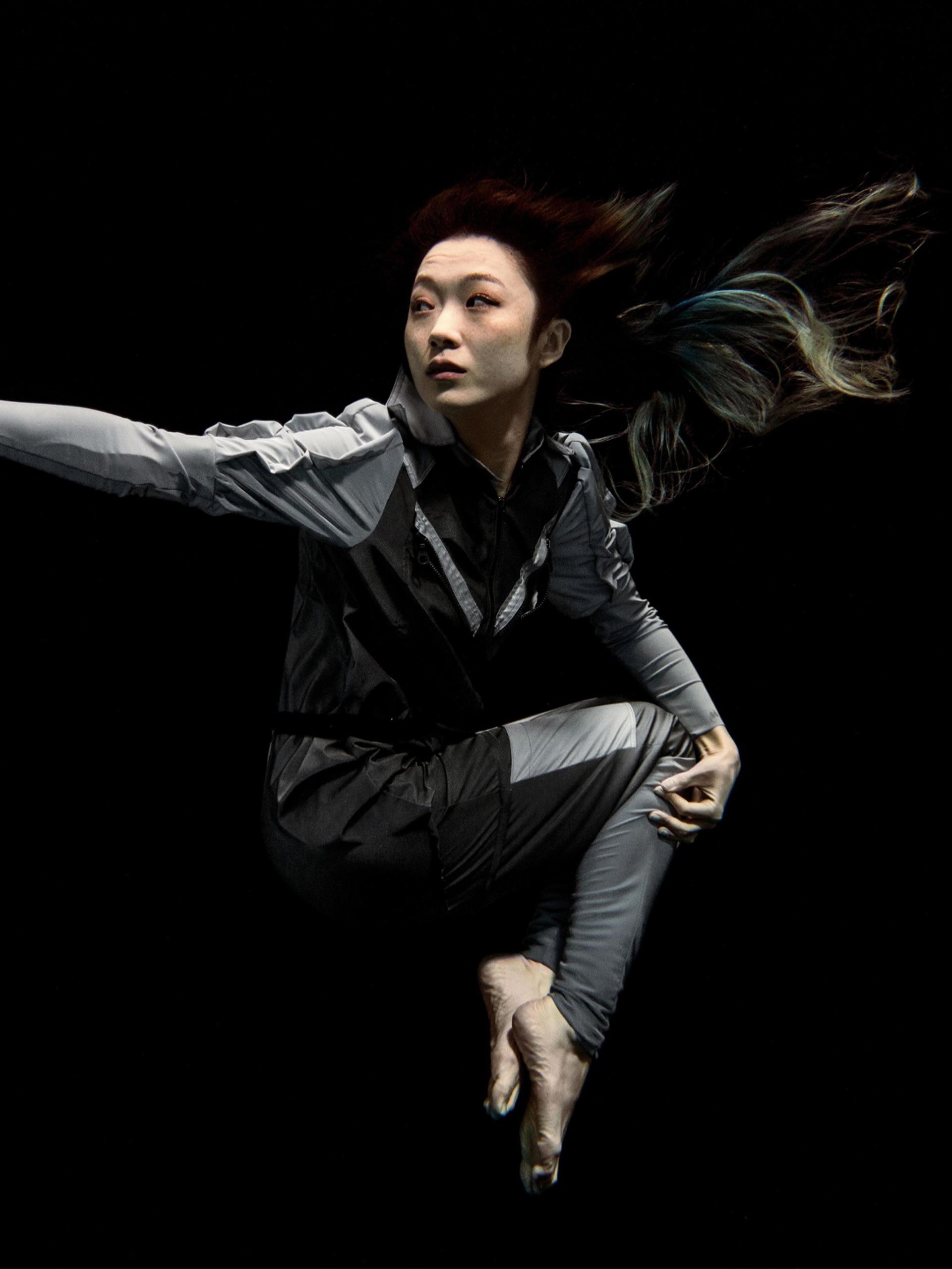 Artist Xin Liu floating with arm outstretched against a black background. She wears a full-length grey body suit with long sleeves with bare feet and hands..