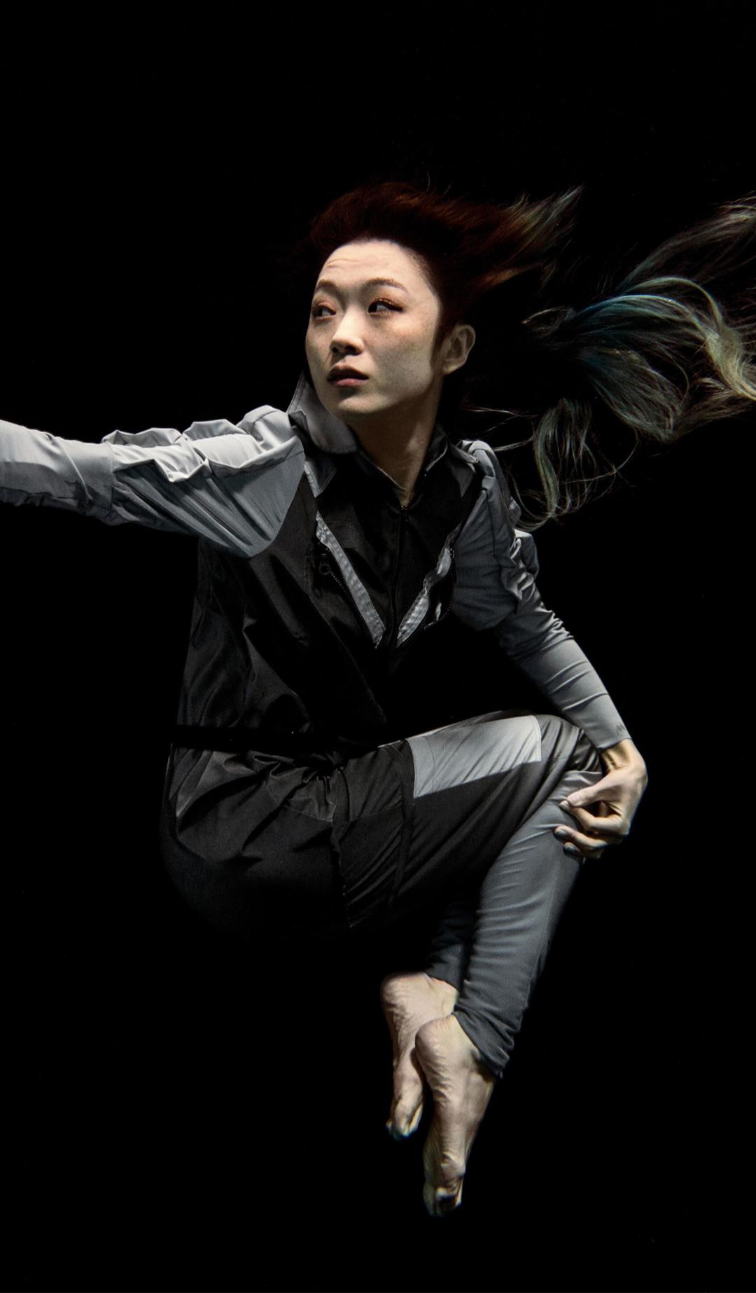 Artist Xin Liu floating with arm outstretched against a black background. She wears a full-length grey body suit with long sleeves with bare feet and hands..