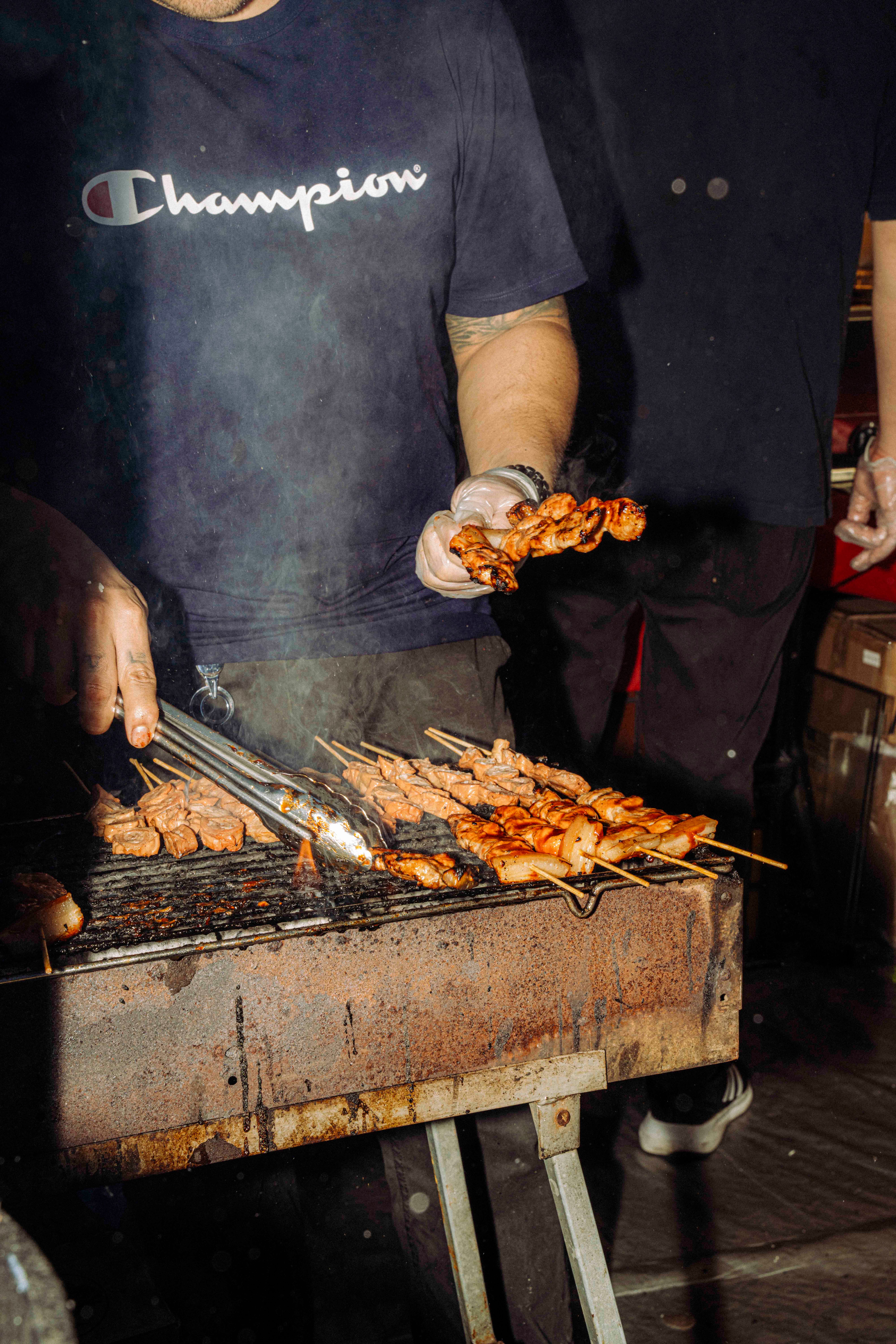 A vendor in a blue ‘Champion’ T-shirt grills skewers of meat over an open flame, holding one skewer while managing another on the grill with a pair of tongs.