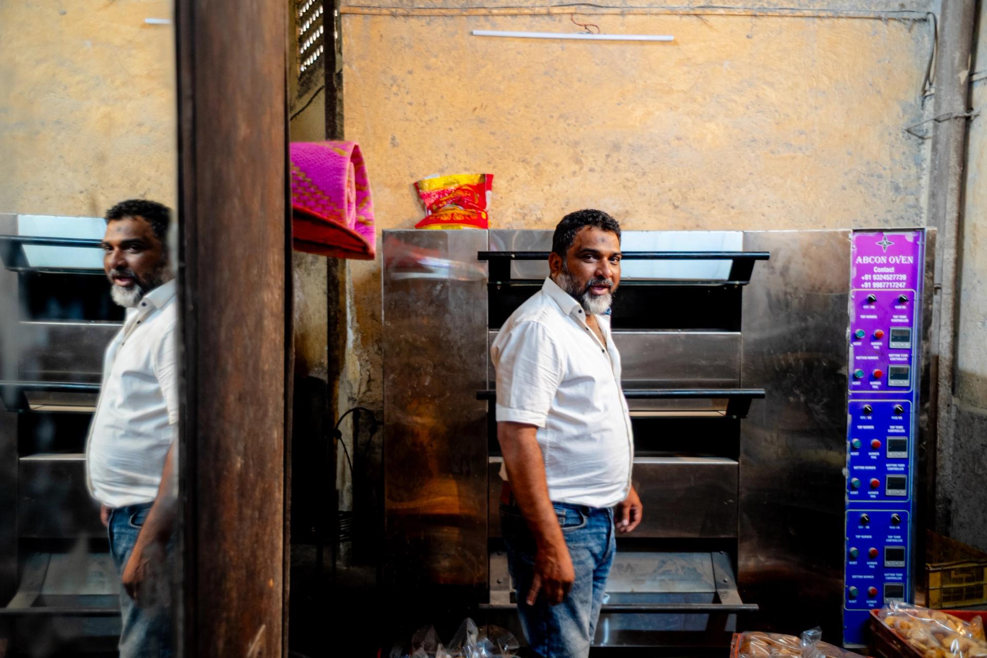 A man in a bakery stands in front of a non-operational gas oven. He wears a short sleeve button-up shirt and jeans.