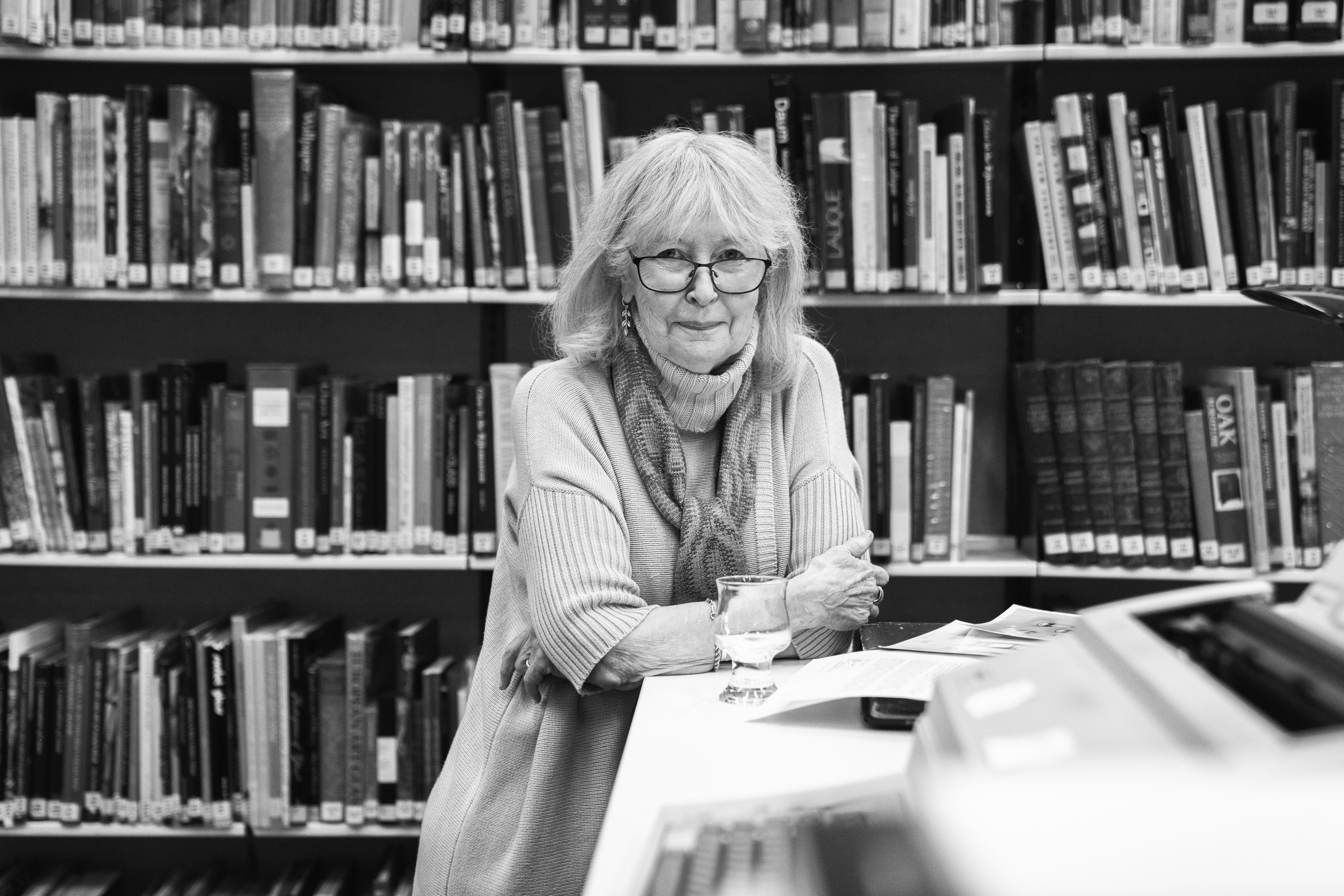 Black and white portrait of woman (Denise Larcombe) in a library, sitting on a table with a curved glass in front of her.