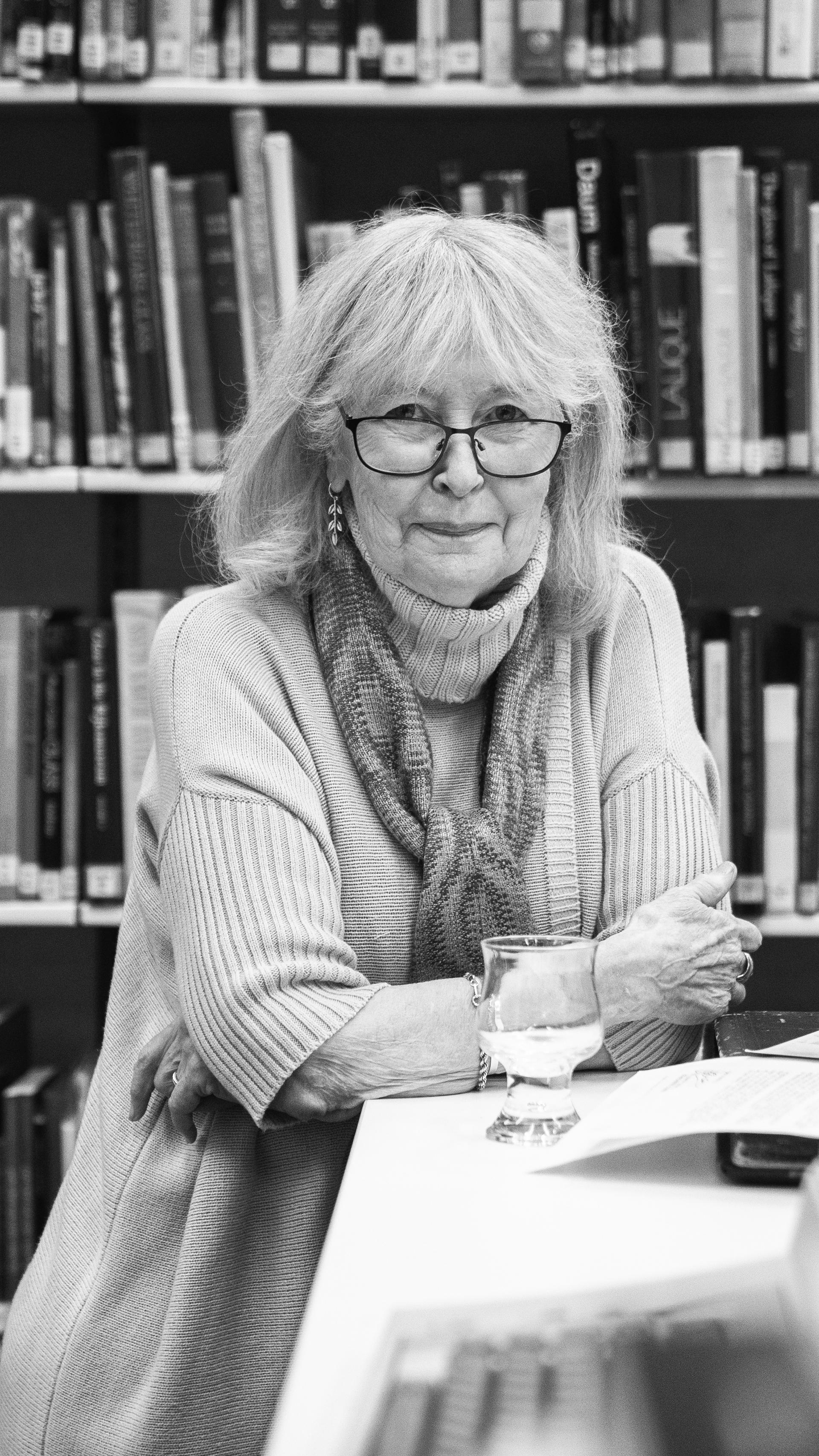 Black and white portrait of woman (Denise Larcombe) in a library, sitting on a table with a curved glass in front of her.