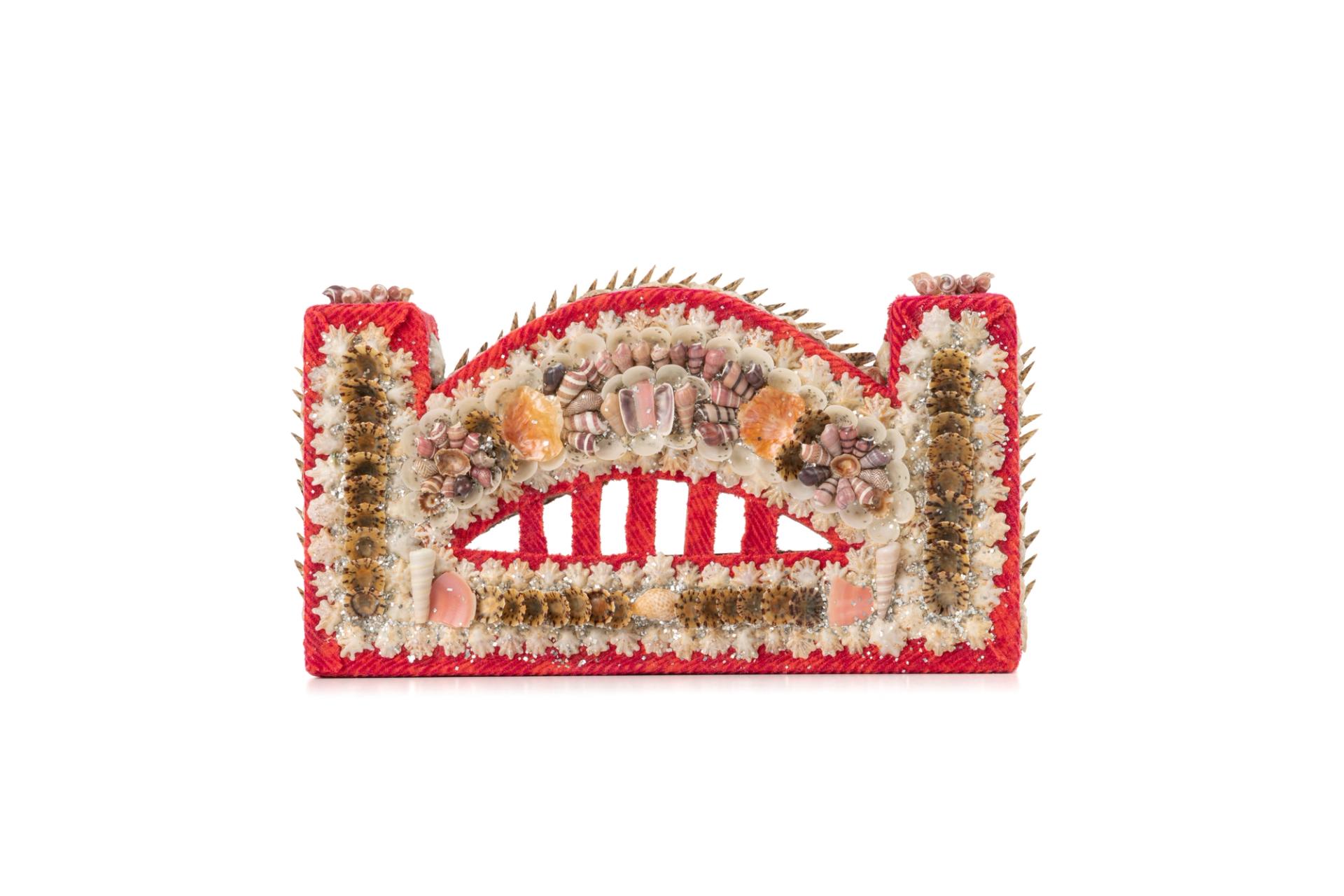 A Harbour Bridge miniature, made of stiff cardboard and covered in covered in orange and red corduroy and decorated with shells arranged in different patterns around the image of the the bridge. There are various types of shells clustered in an abstract design to create a decorative facade.