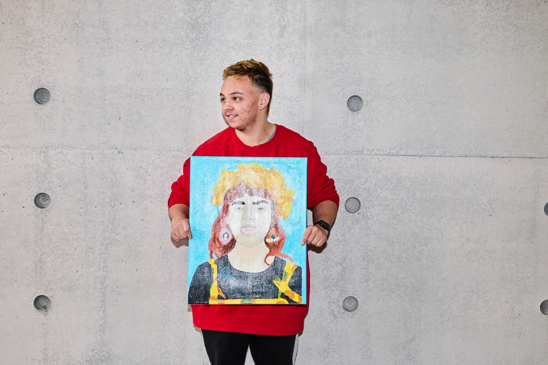 A guest in a red jumper, holding a painted portrait of Chloe Hayden against a concrete wall.