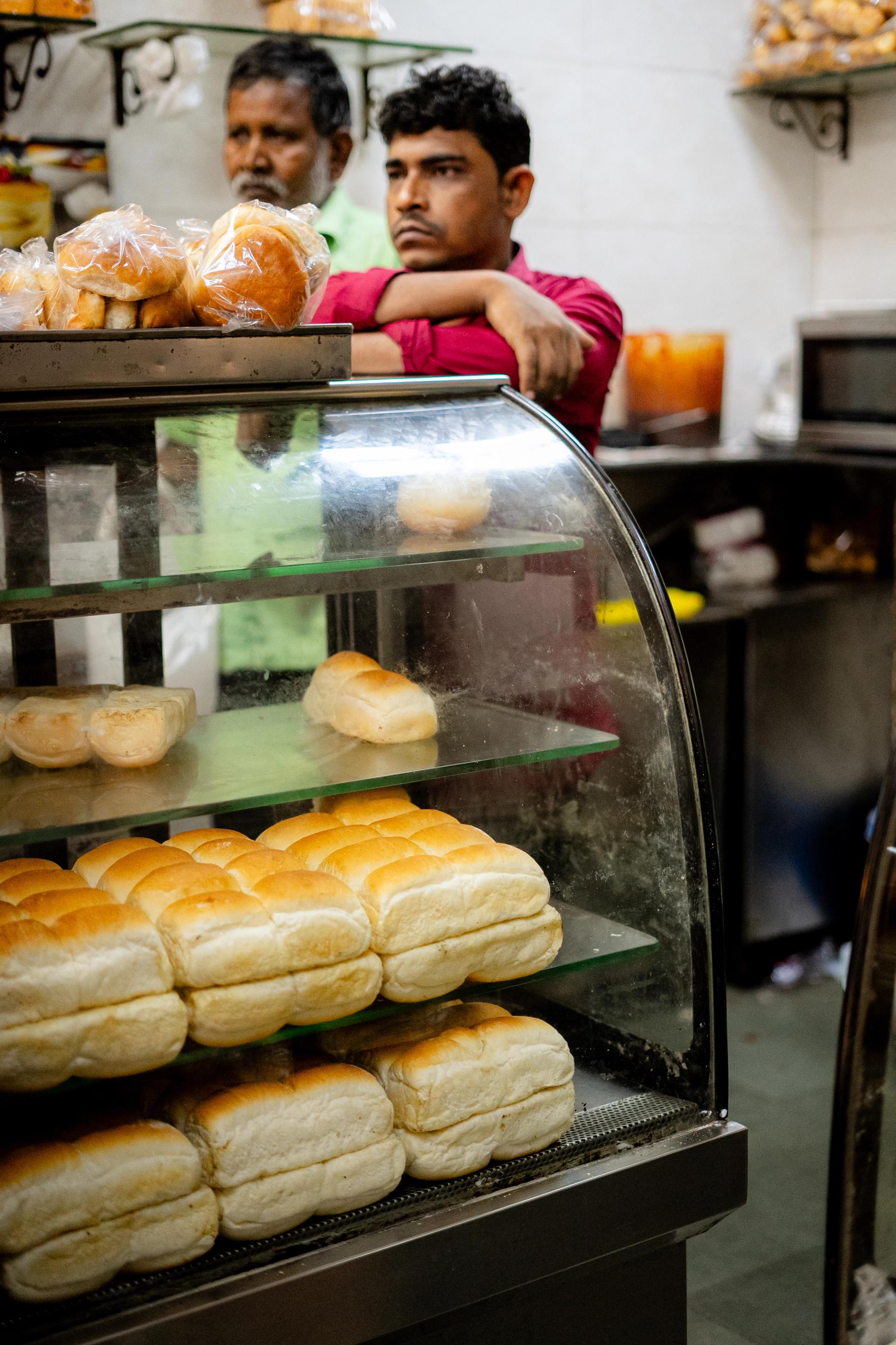 Two men in a bakery behind a glass cabinet filled with bread rolls. One man wears a red button-up shirt and rests his crossed arms on the back of the cabinet.