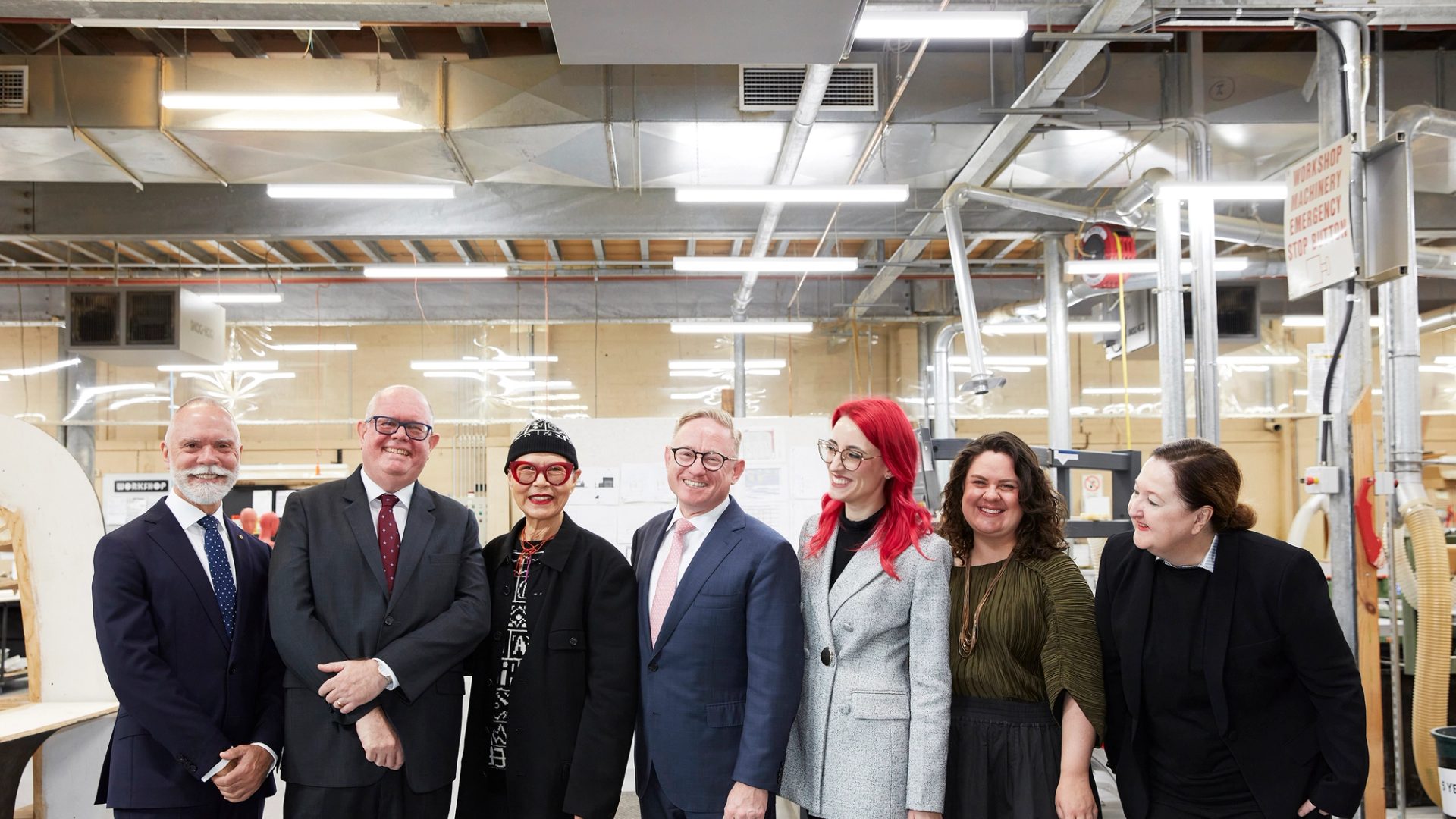 A group of people pose for a photo standing in a line, they smile at the camera and each other. They are in a workshop space.