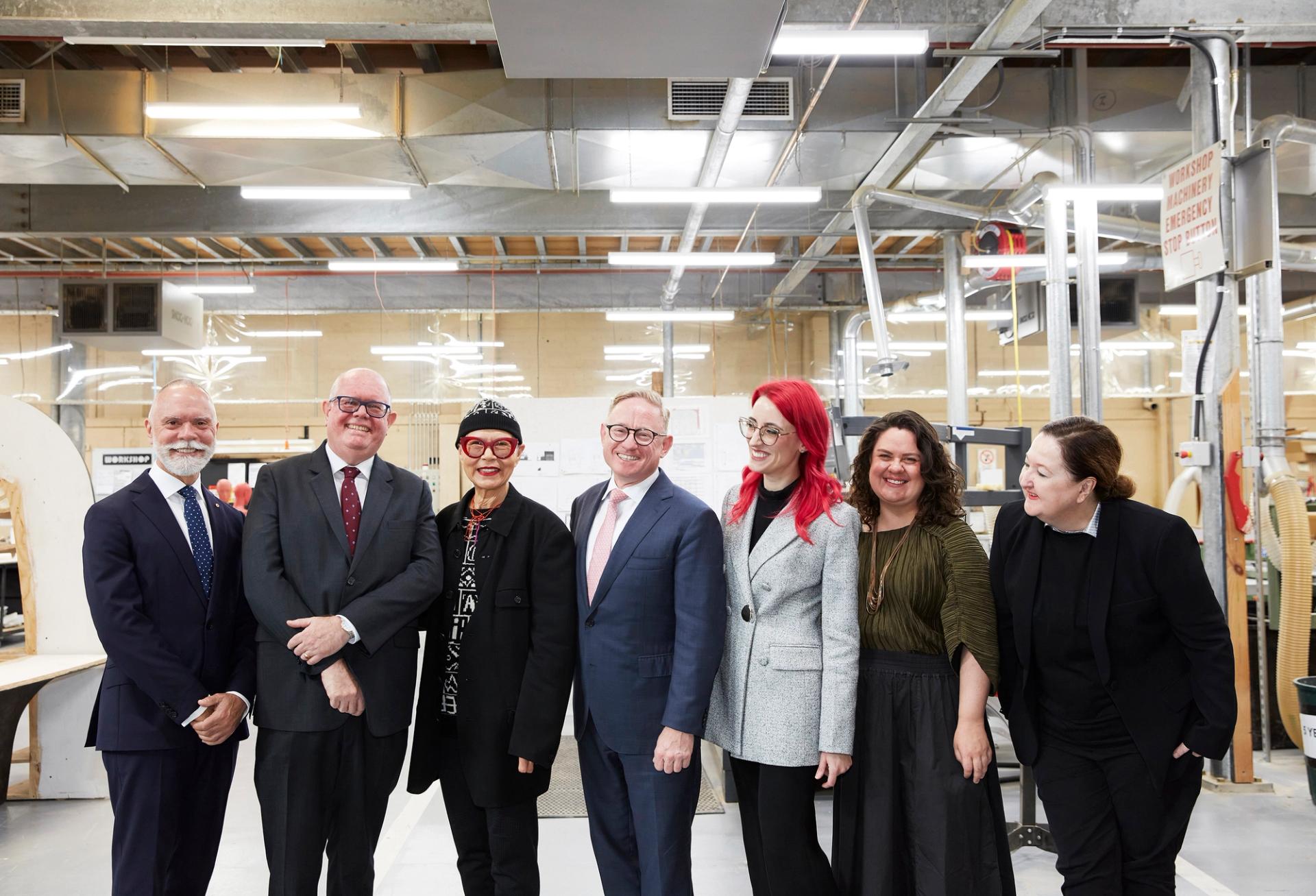 A group of people pose for a photo standing in a line, they smile at the camera and each other. They are in a workshop space.