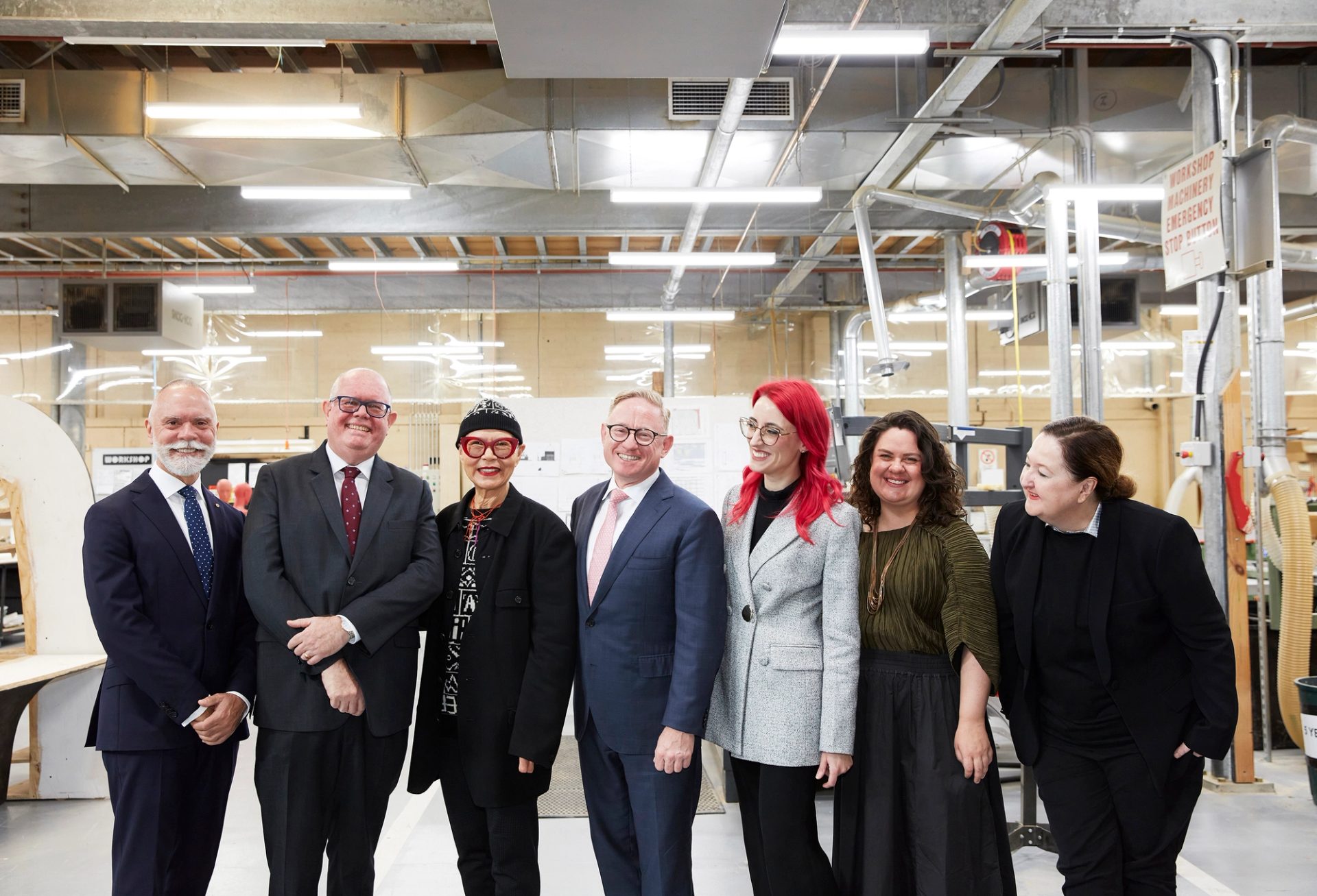 A group of people pose for a photo standing in a line, they smile at the camera and each other. They are in a workshop space.