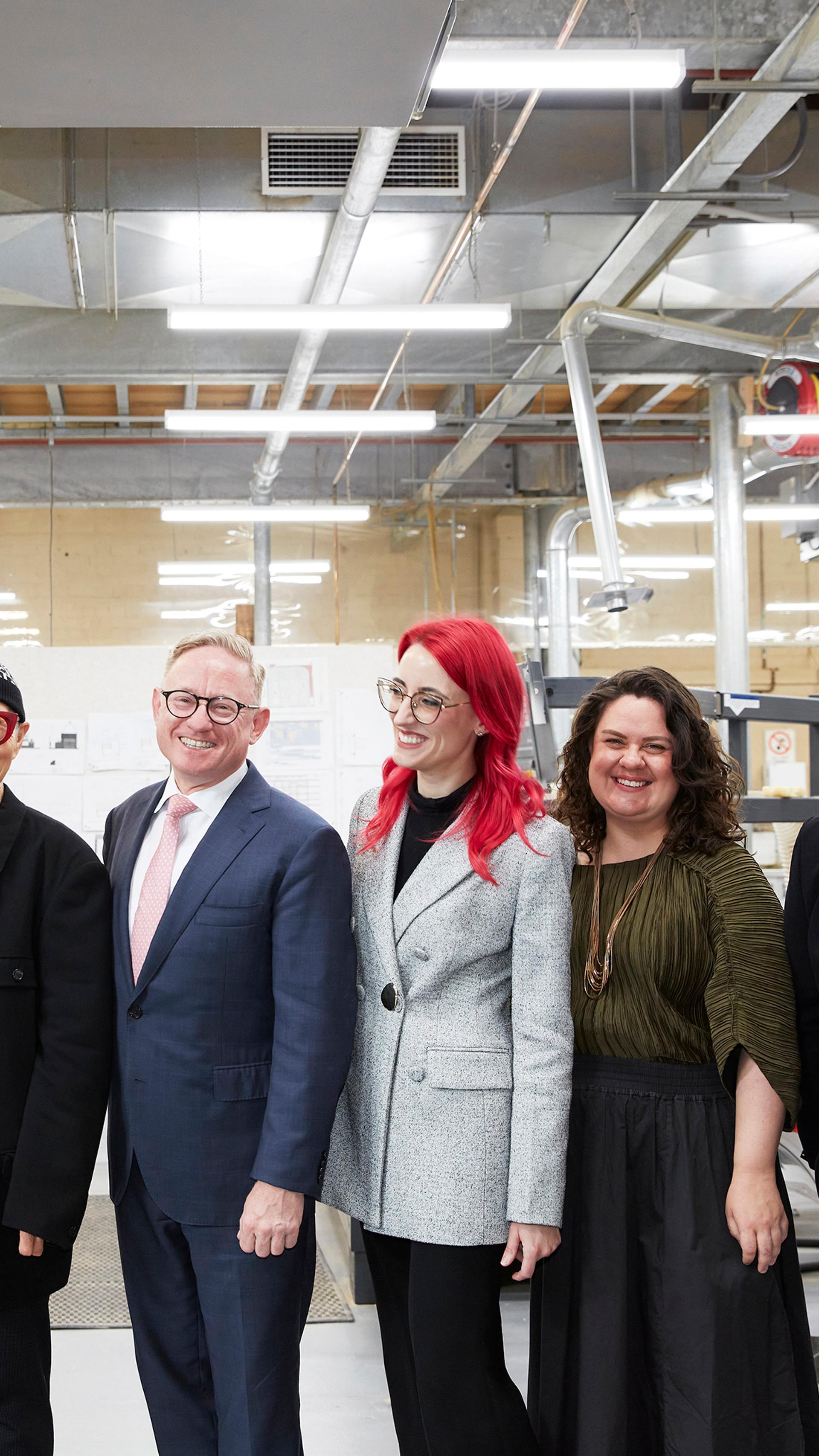 A group of people pose for a photo standing in a line, they smile at the camera and each other. They are in a workshop space.