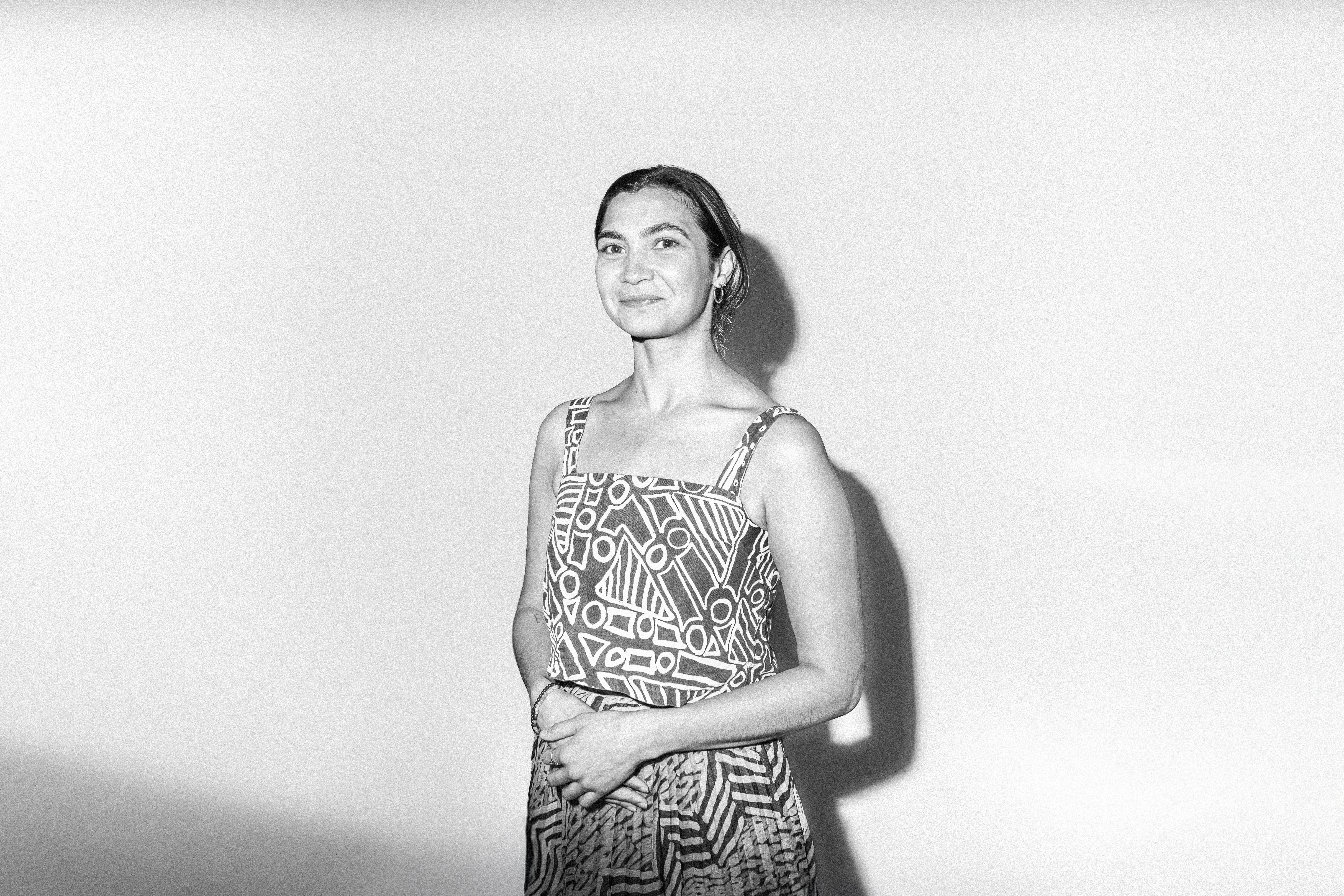 A woman with short hair wearing a strap sleeve shirt with intricate patterns (Rachael Hocking) stands against a white background, with the cameras flash casting a shadow. They are facing the camera with their hands are held together at her waist.