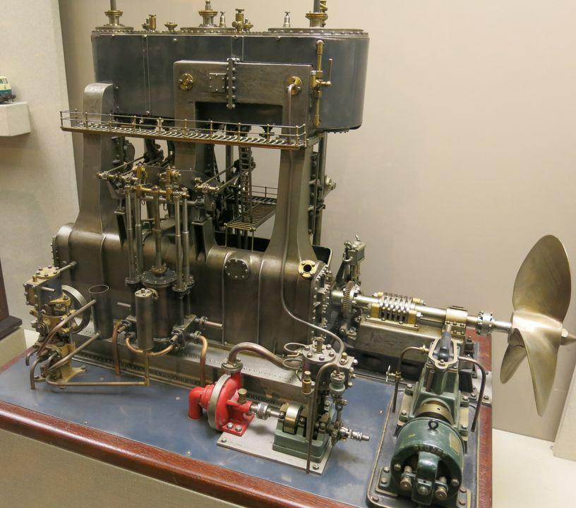 Small silver metal engine model with propeller on the right side bolted to a bench