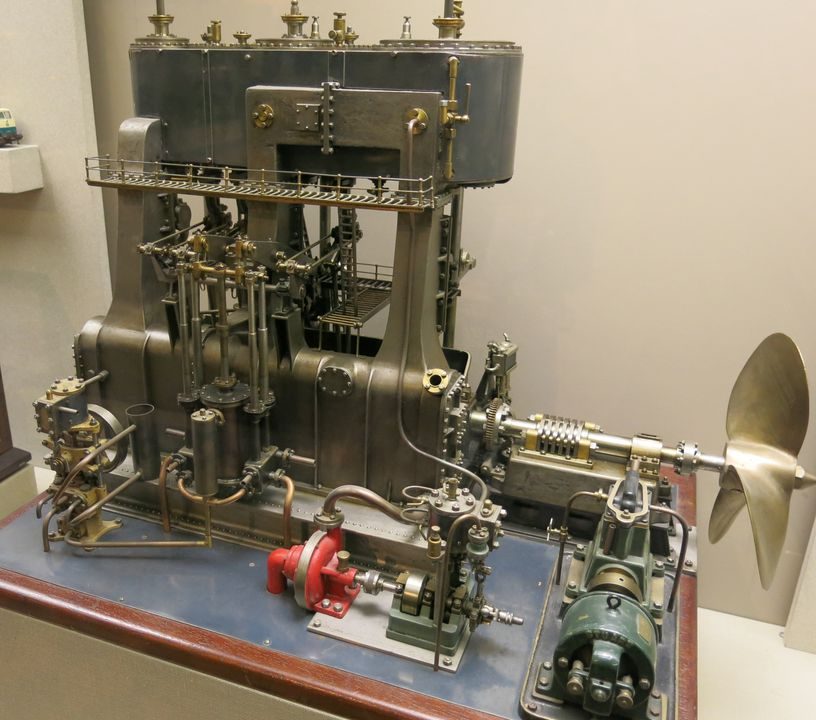 Small silver metal engine model with propeller on the right side bolted to a bench