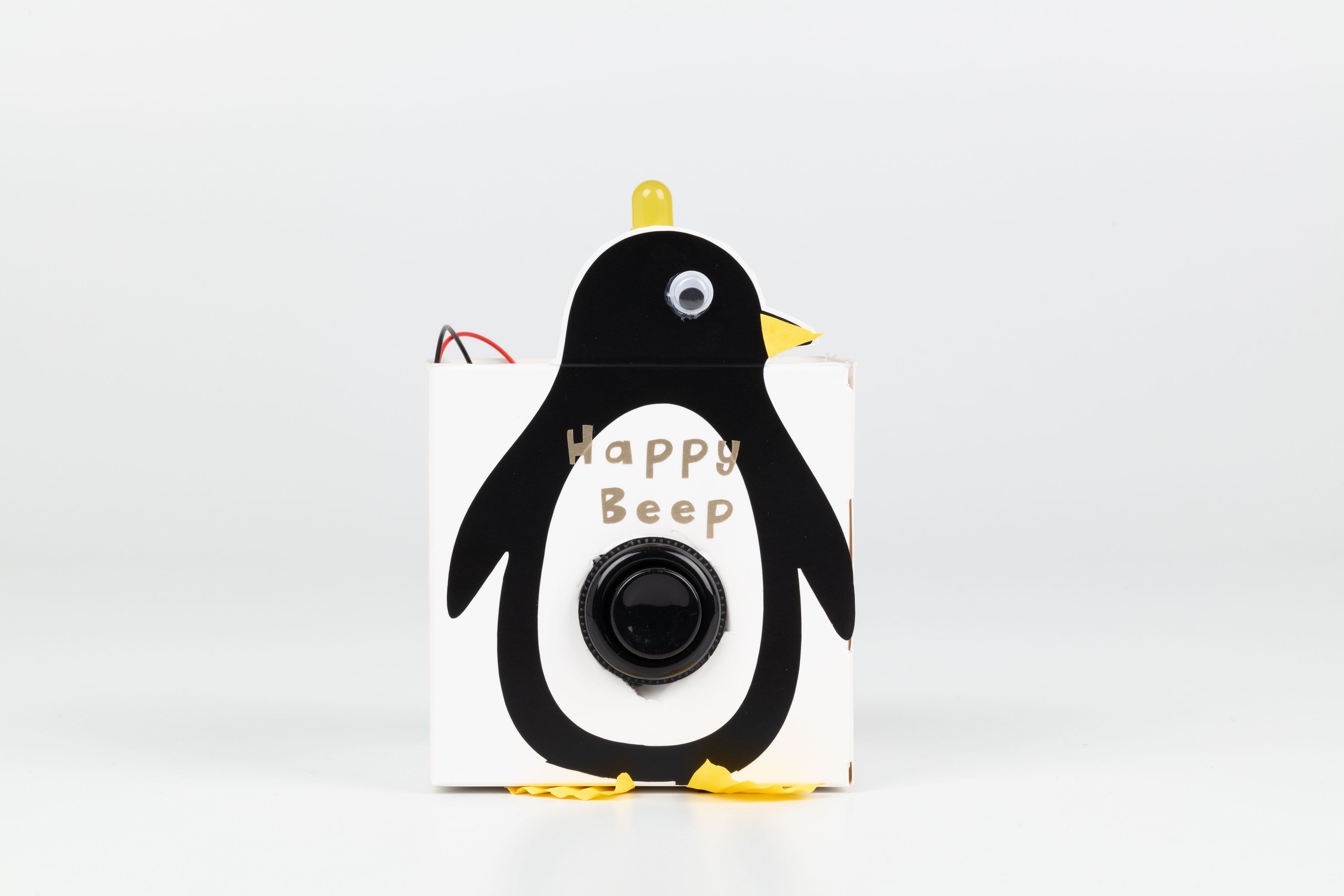 Image of a hand crafted mechanical device against a white background. A box with a cutout of a penguin and a button in the centre, the words ‘Happy Beep’ above the button. A yellow light on the top of the penguin’s head.