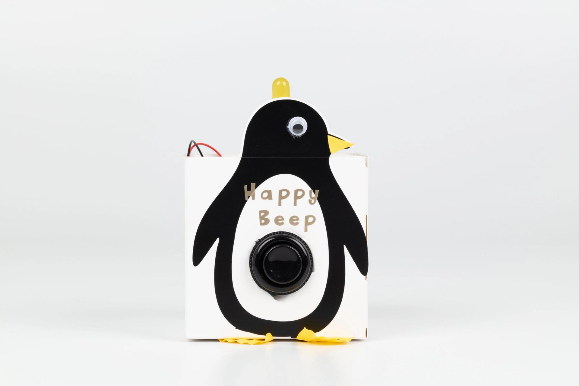 Image of a hand crafted mechanical device against a white background. A box with a cutout of a penguin and a button in the centre, the words ‘Happy Beep’ above the button. A yellow light on the top of the penguin’s head.